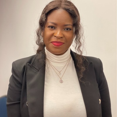 Smiling woman with curly hair wearing red lipstick, a white turtleneck, layered necklaces, and a black blazer.