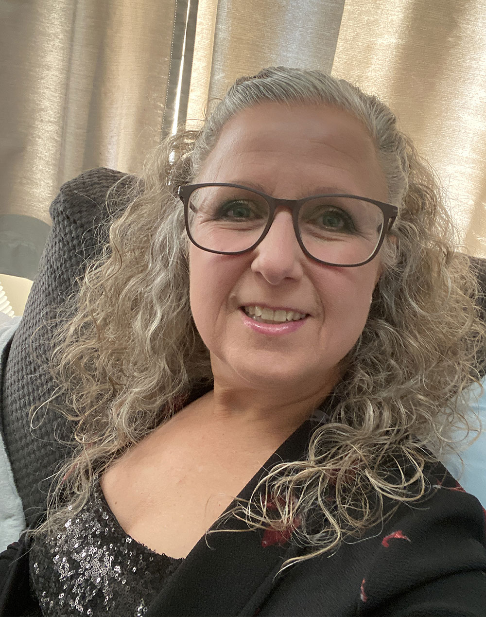 Smiling woman with curly blonde hair and glasses wearing a black sequined top and jacket.