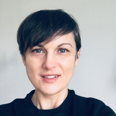 Woman with short dark hair and a black top smiling against a plain light background.