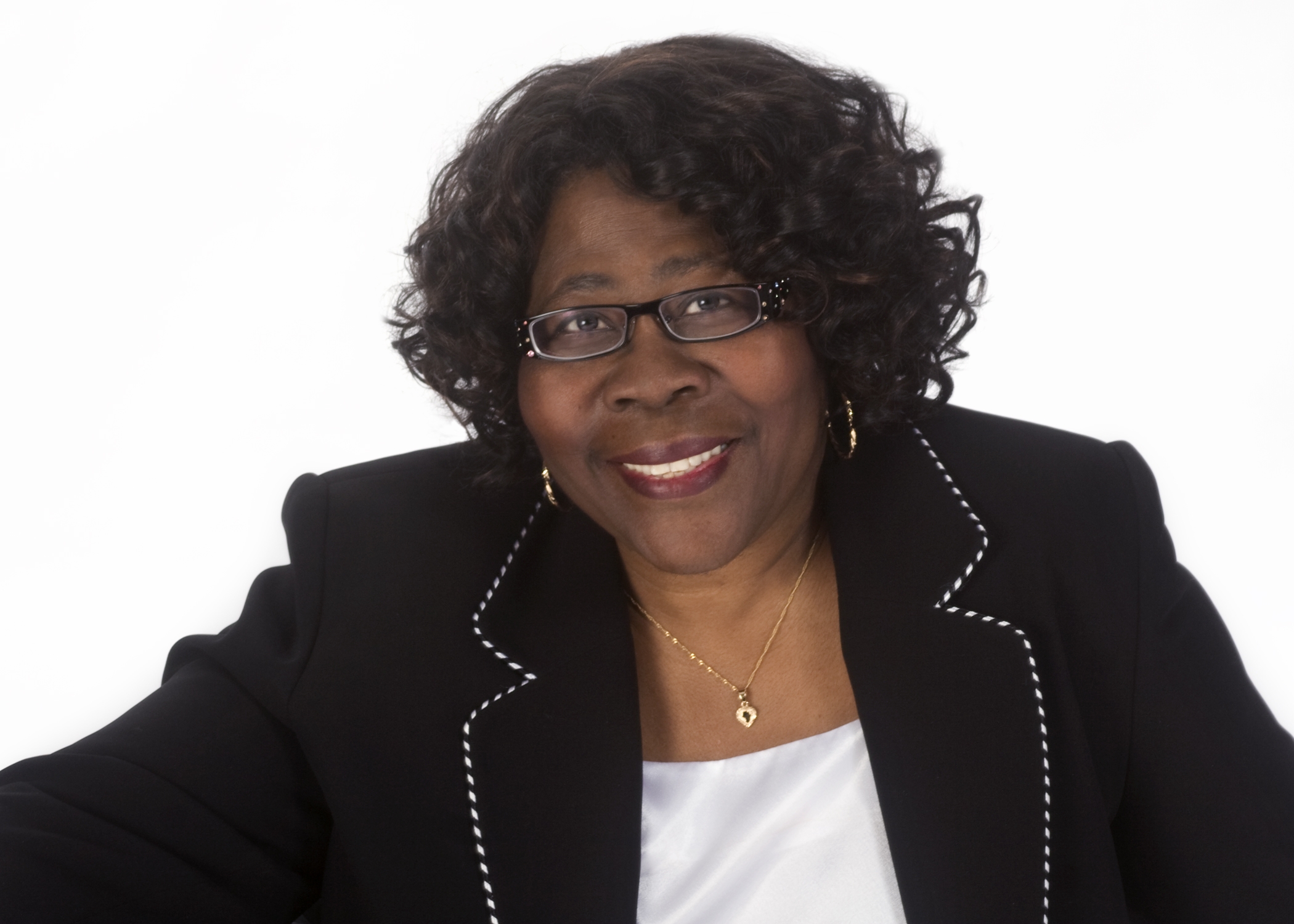 Smiling woman with curly hair wearing glasses, gold jewelry, black blazer with white trim.