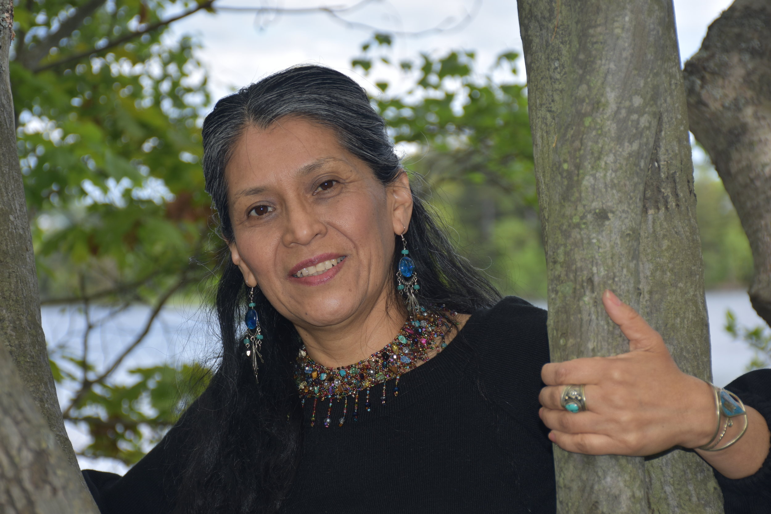 Woman with long dark hair smiling, wearing colorful jewelry, standing between two tree trunks outdoors.