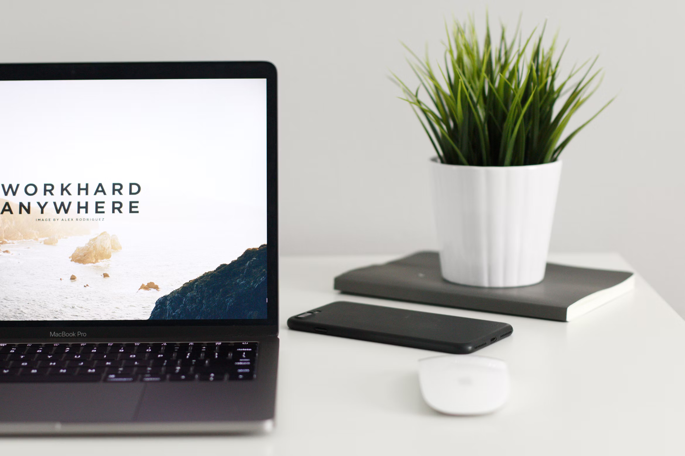 Laptop with 'Work Hard Anywhere' on screen, smartphone, white mouse, and a plant on a clean desk.