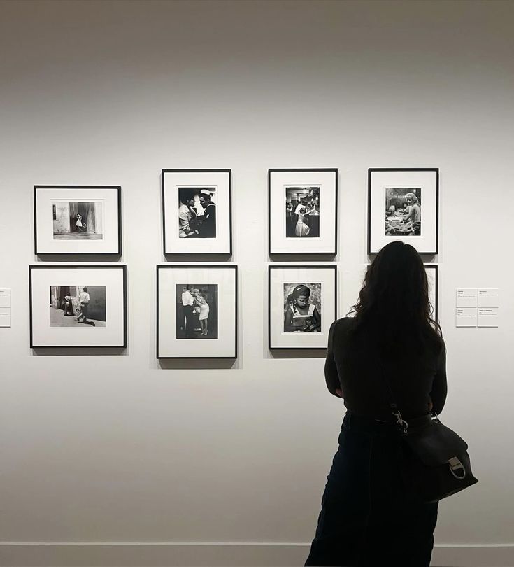 Person with long hair viewing eight framed black-and-white photos on a white gallery wall.