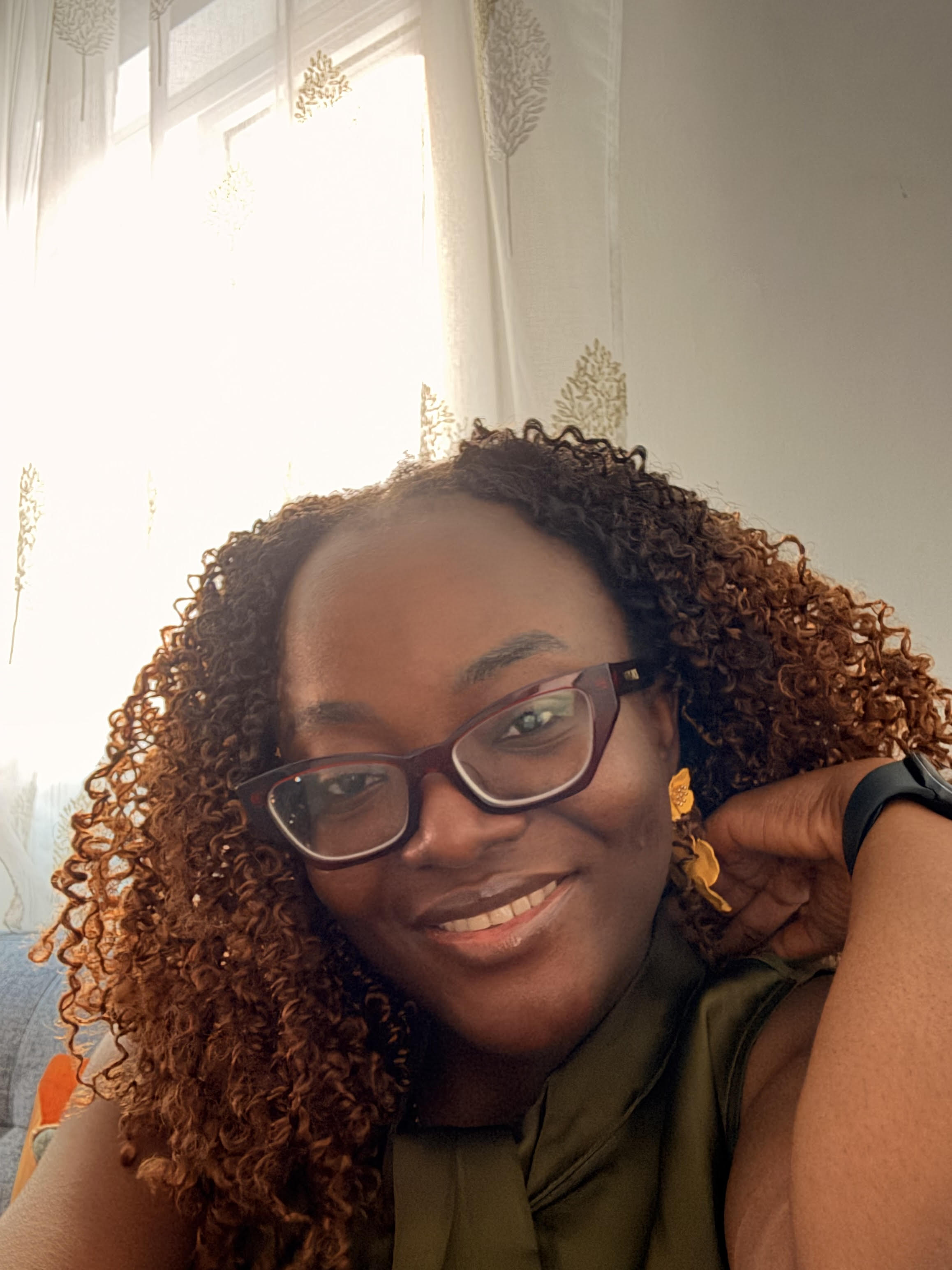 Smiling woman with curly hair, glasses, yellow earrings, and a green top near a window with sheer curtains.
