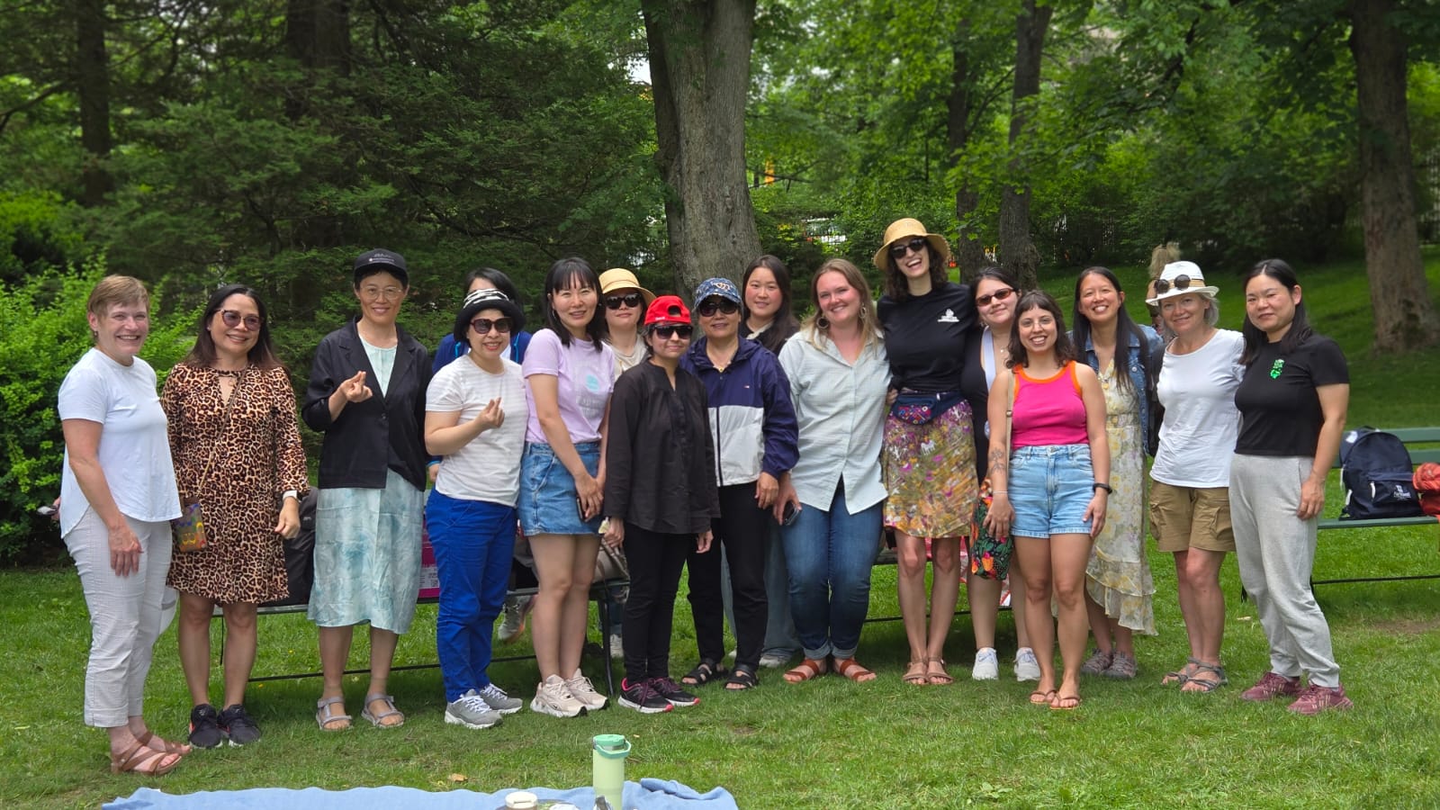 Group of diverse women standing on grass in a park with trees in the background.