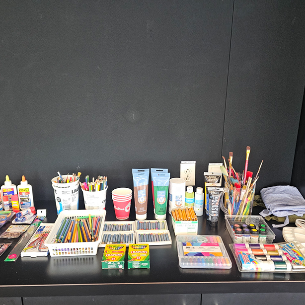 Table with various art supplies including paints, brushes, crayons, colored pencils, and glue bottles.