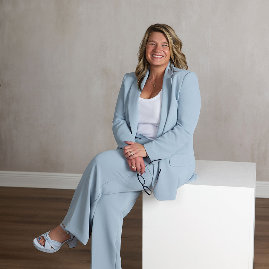 Women’s leadership coach seated in a light blue suit, smiling and holding glasses in a professional studio setting.