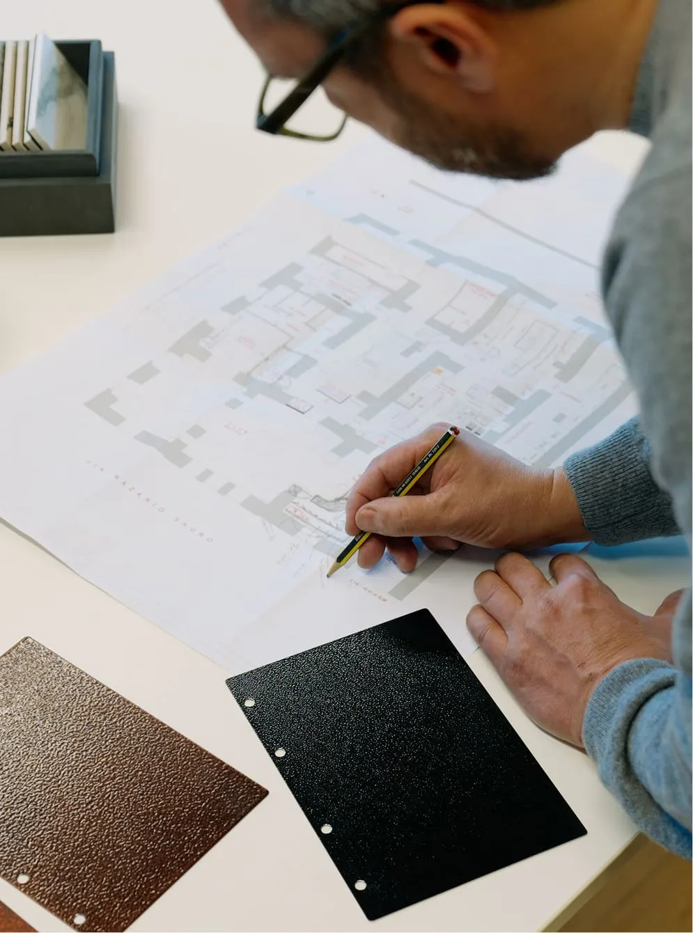 Man wearing glasses and gray sweater making notes on an architectural floor plan with two textured material samples on the table.
