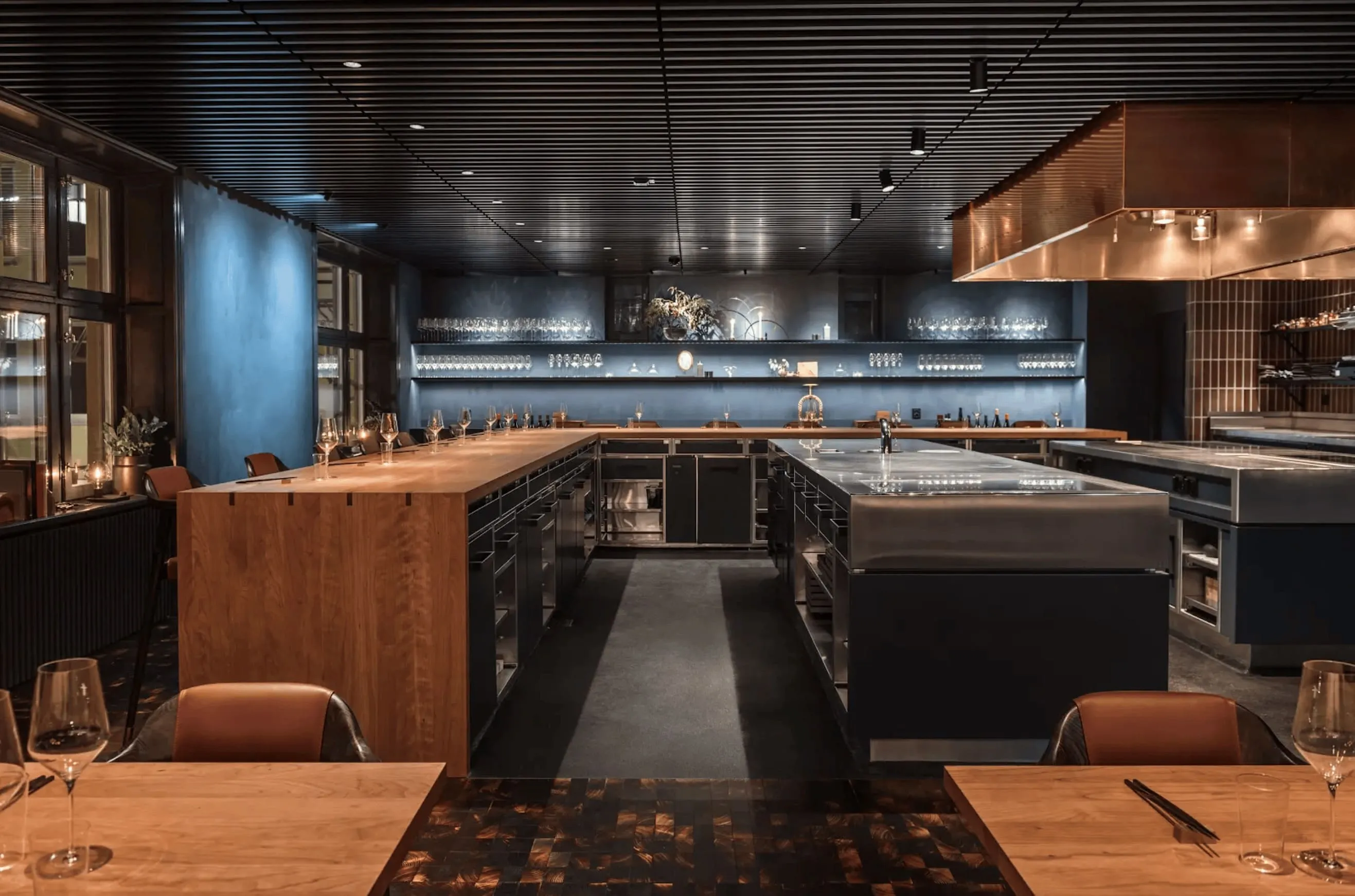 Modern restaurant kitchen with large cooking islands, wooden counters, and shelves of glassware against blue walls.