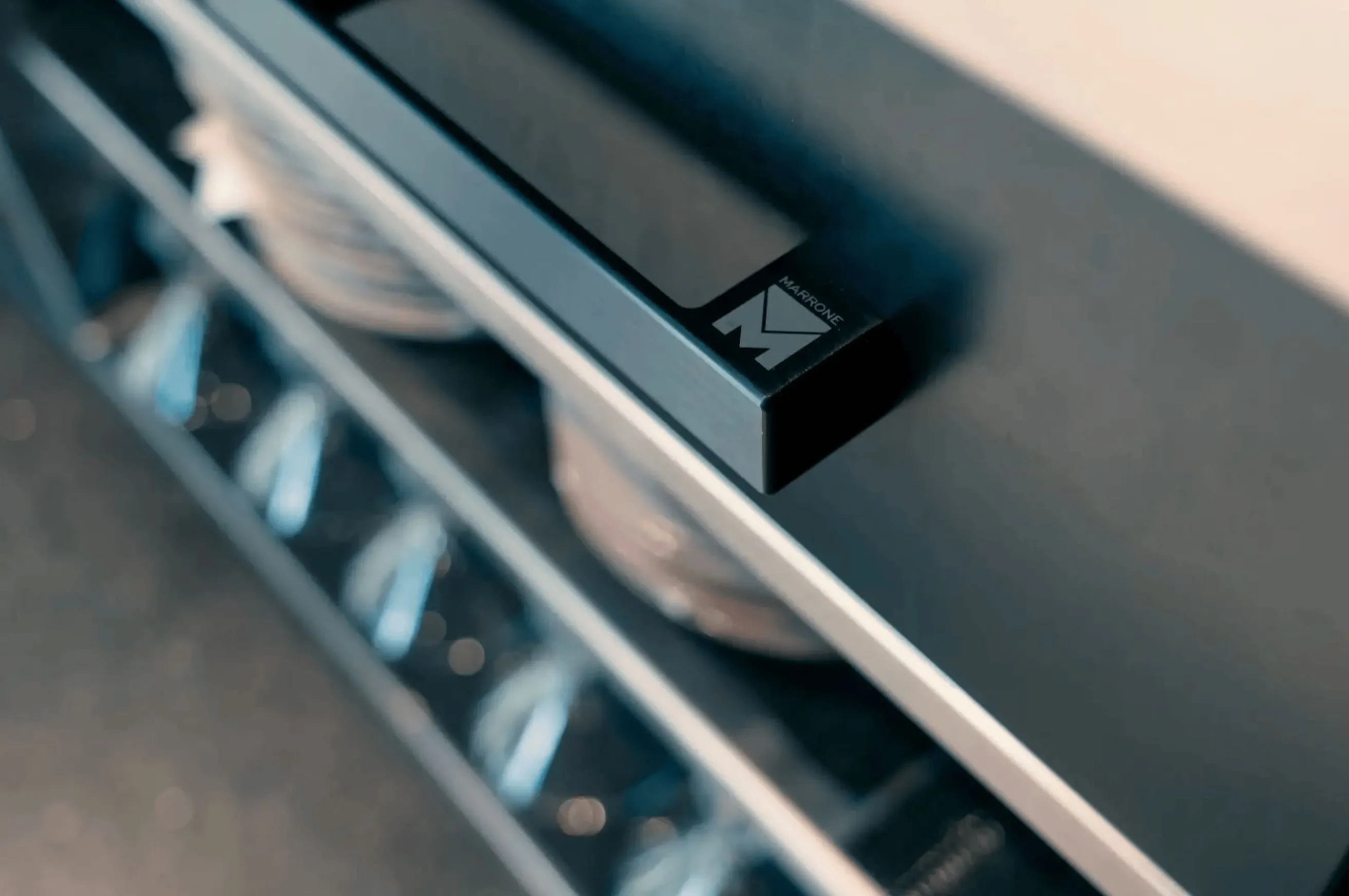 Close-up of a black cabinet handle with the brand name Marrone and the letter M on it, with blurred shelves and glassware in the background.