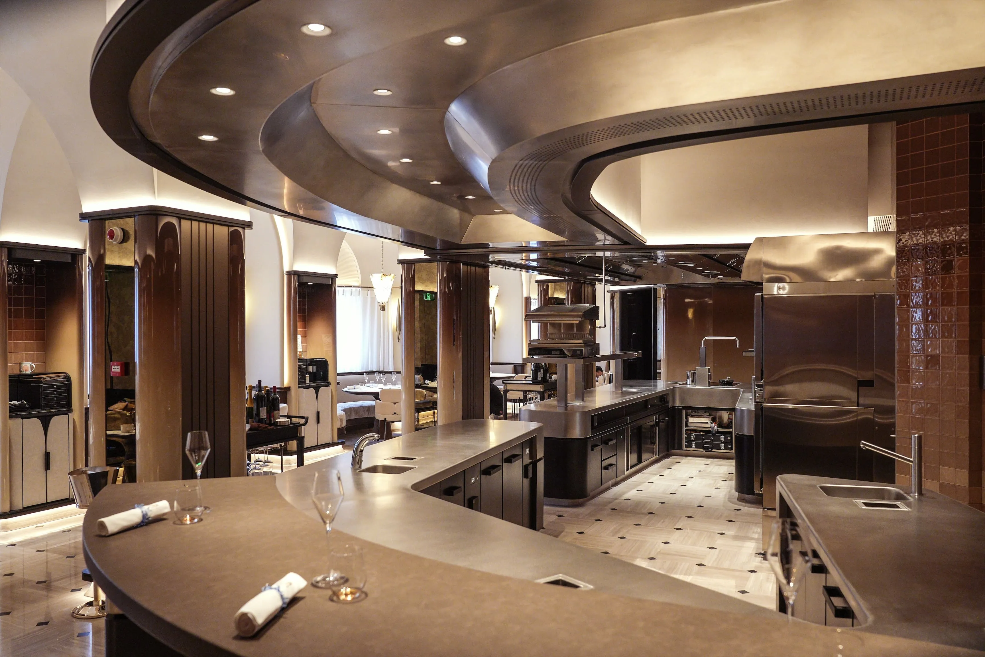 Modern, elegant restaurant interior with curved bar counters, stainless steel kitchen equipment, and ambient lighting.