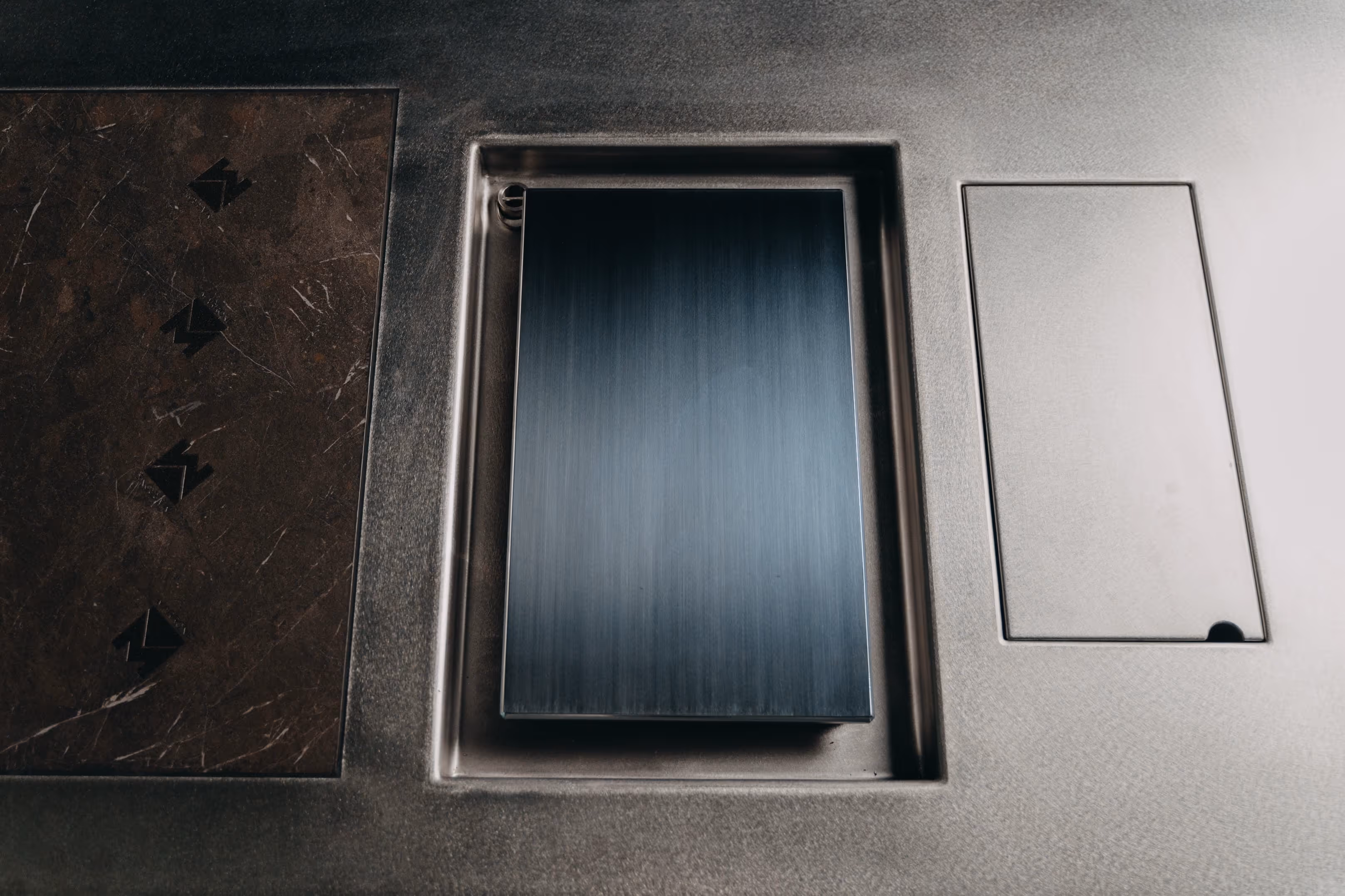 Close-up of three built-in kitchen components with a dark marble panel, a brushed metal flip-up outlet cover, and a small rectangular access panel.