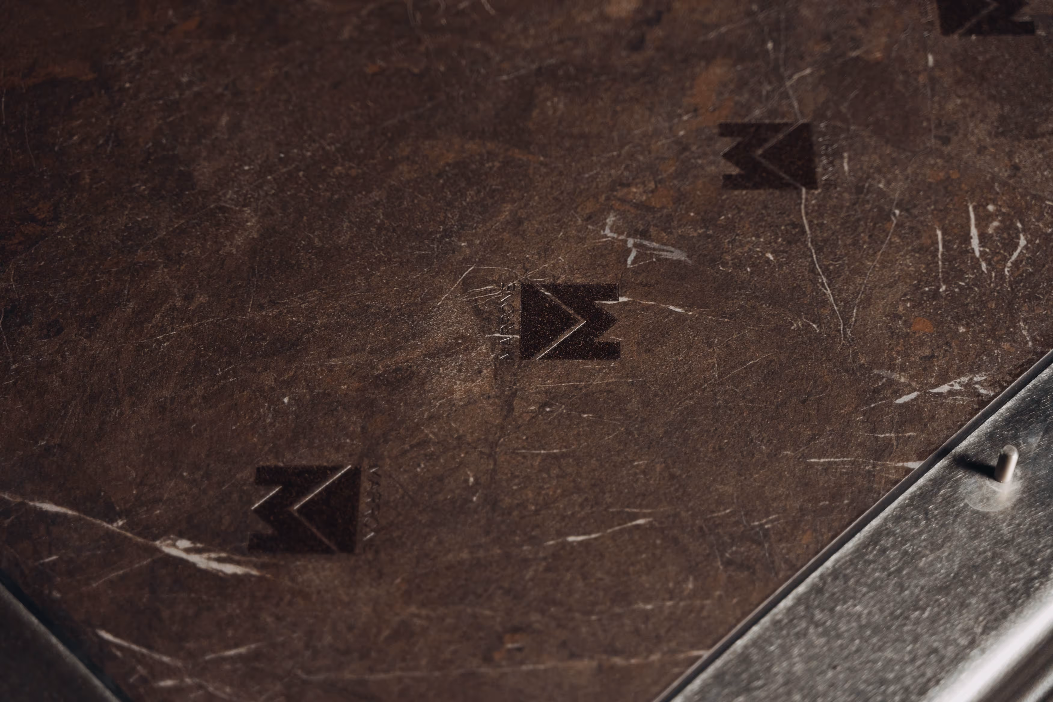 Close-up of a scratched brown surface with three imprinted black M-shaped logos and a metallic edge with a small peg.