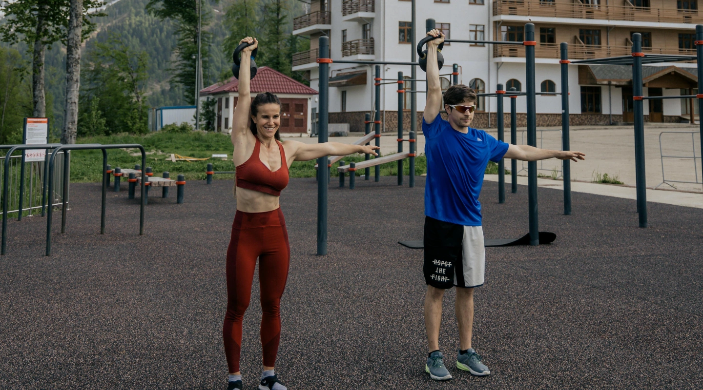 Two people exercise with kettlebells on an outdoor gym, set against a backdrop of a large building and snow-capped mountains under a cloudy sky.