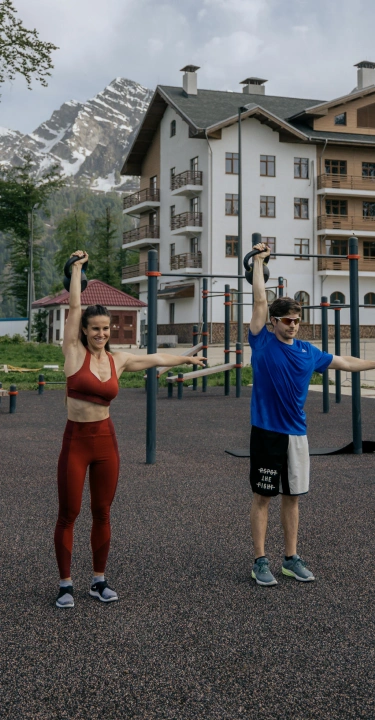 Two people exercise with kettlebells on an outdoor gym, set against a backdrop of a large building and snow-capped mountains under a cloudy sky.