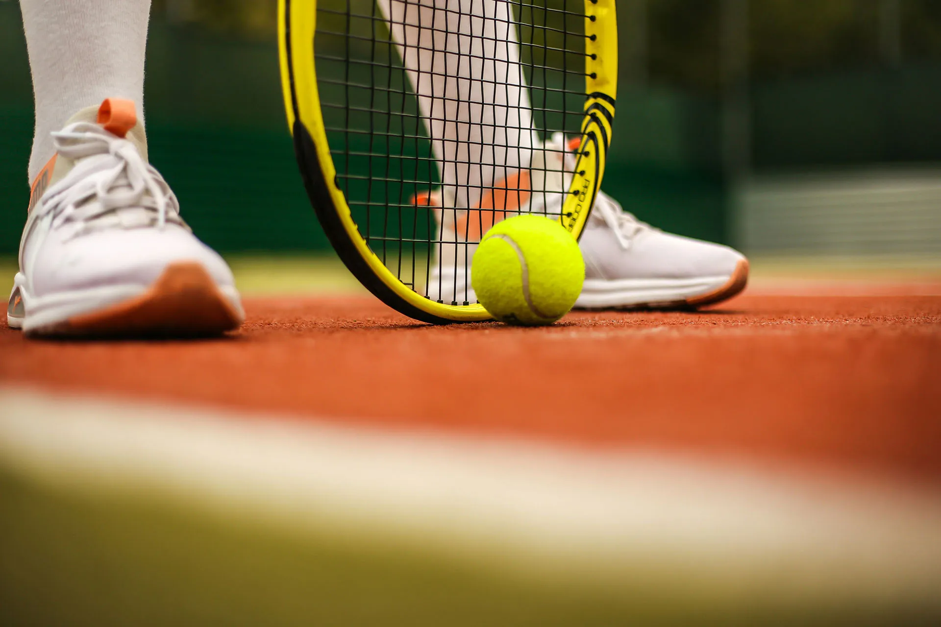 Close-up of a tennis racket touching a yellow tennis ball on a clay court with white shoes nearby.