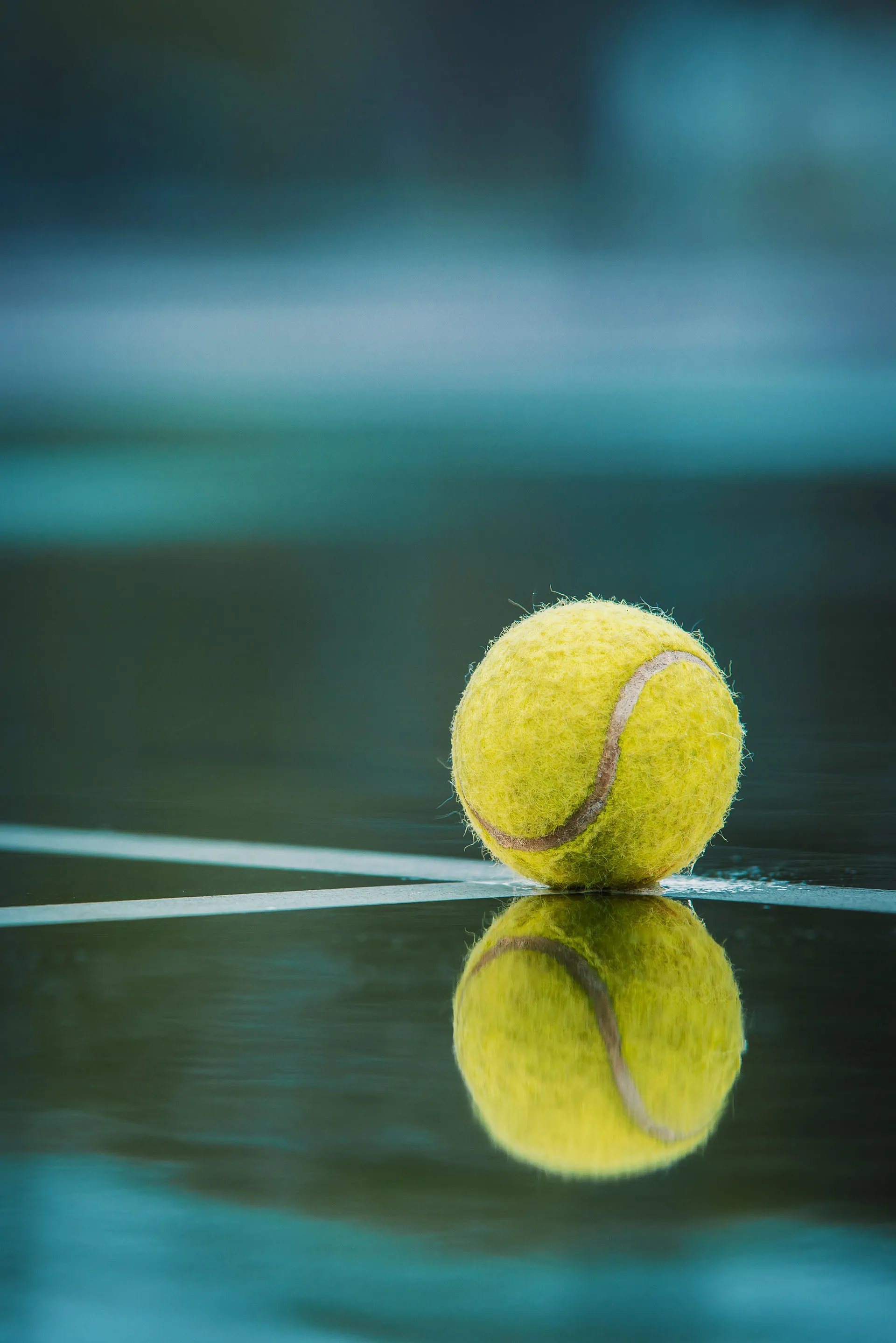 Yellow tennis ball resting on a wet tennis court with its reflection visible on the surface.