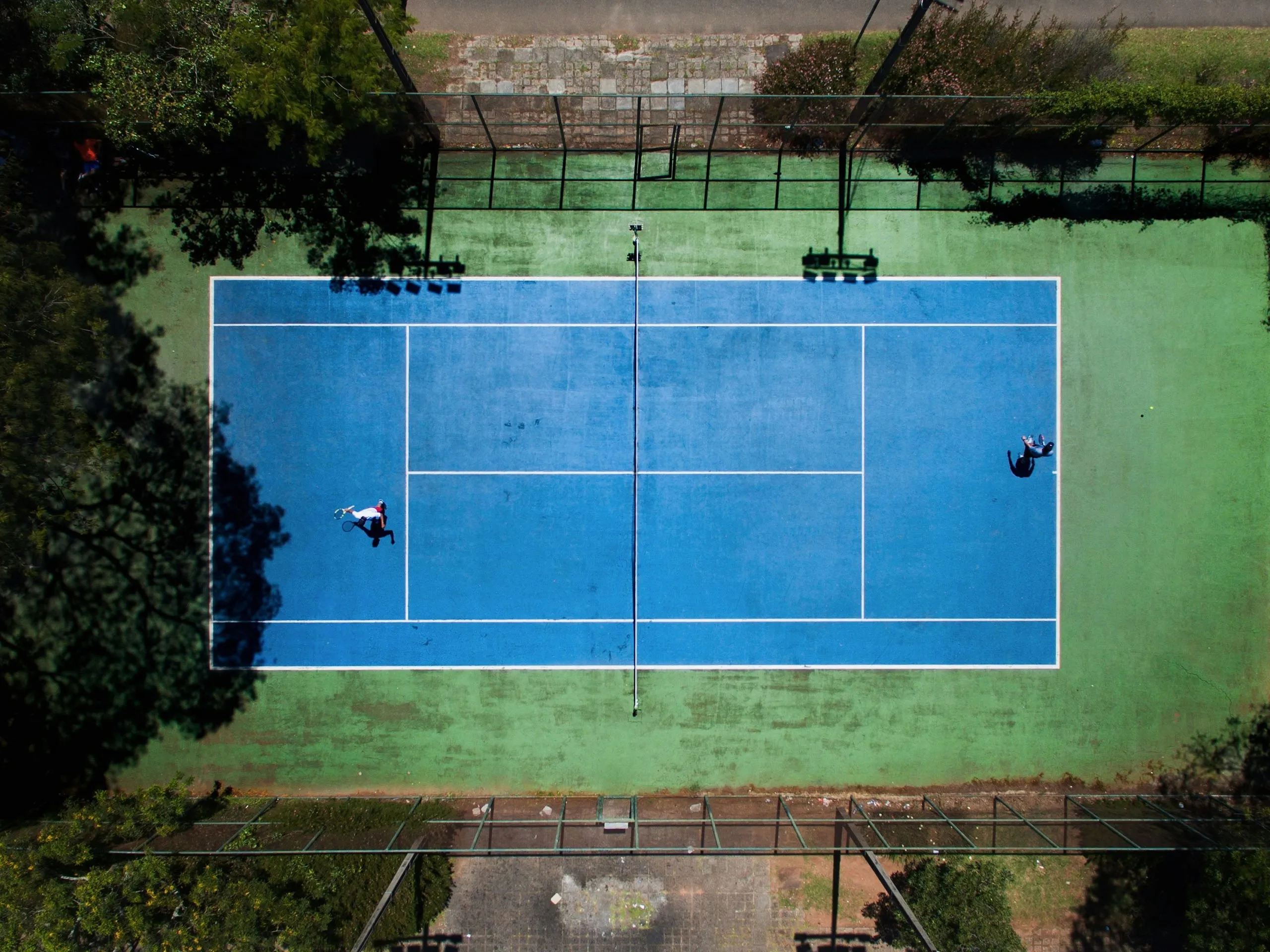 Aerial view of two people playing tennis on a blue court with green surroundings and shadows of nearby trees.