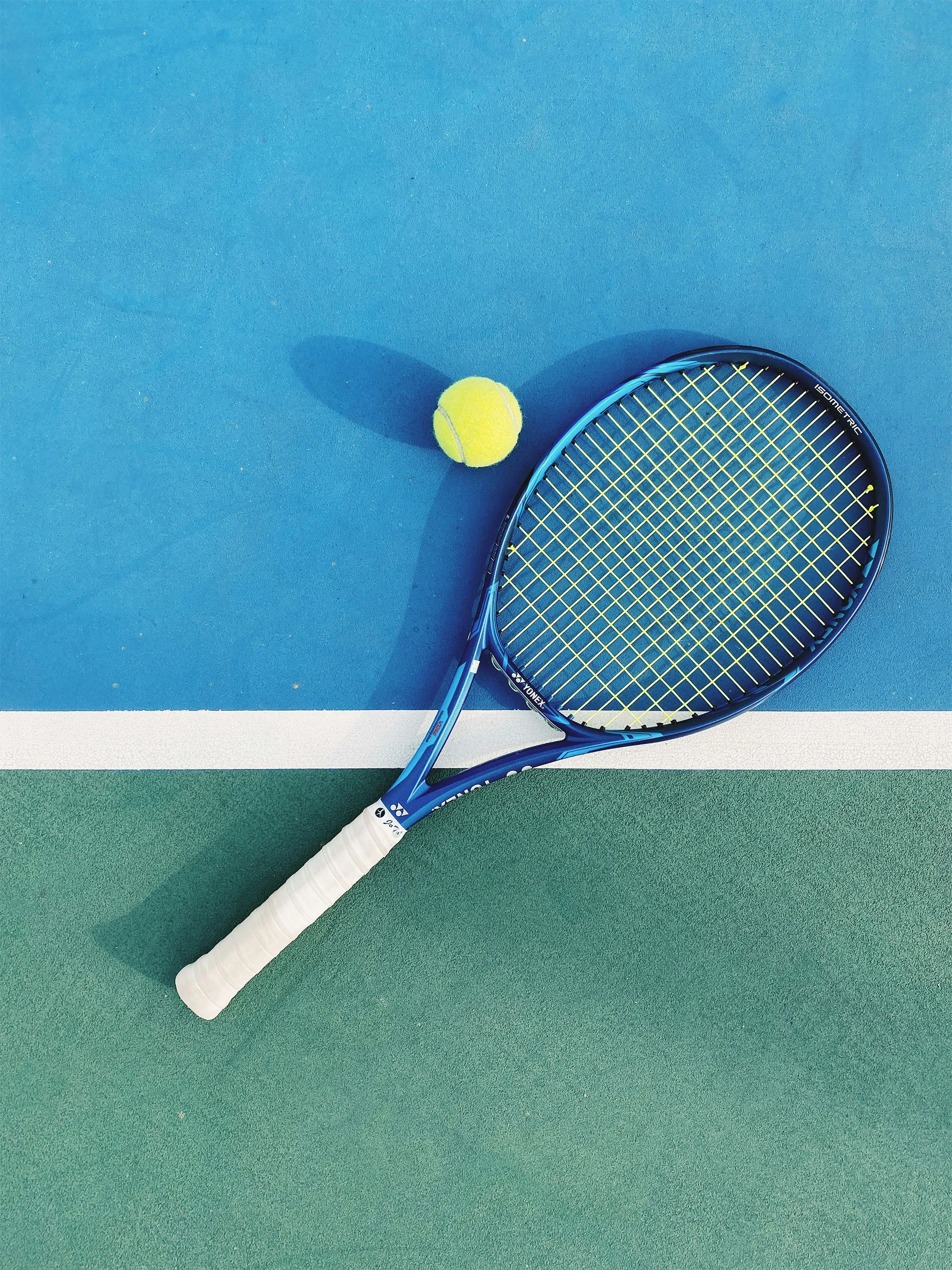 Blue and green tennis court surface with a blue tennis racket and a yellow tennis ball casting shadows.