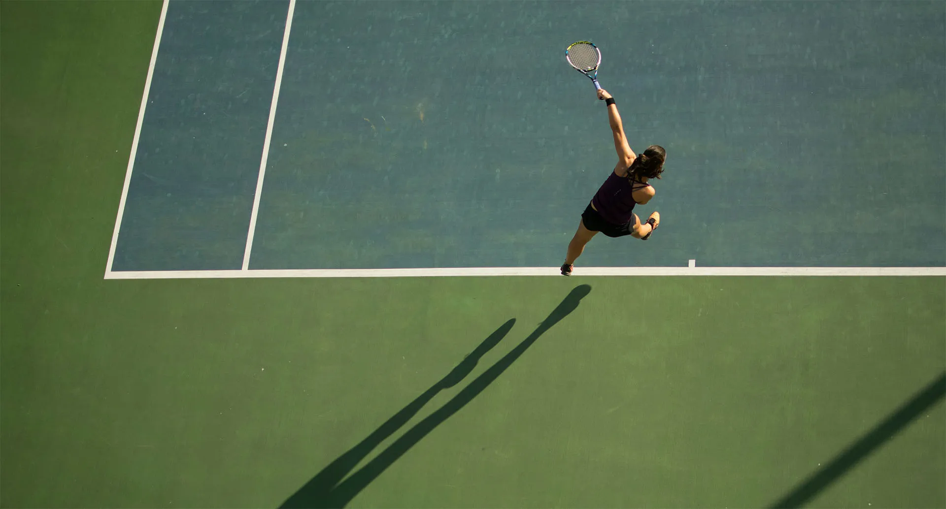 A female tennis player jumping to hit a high forehand shot on a green and blue tennis court from an aerial view.