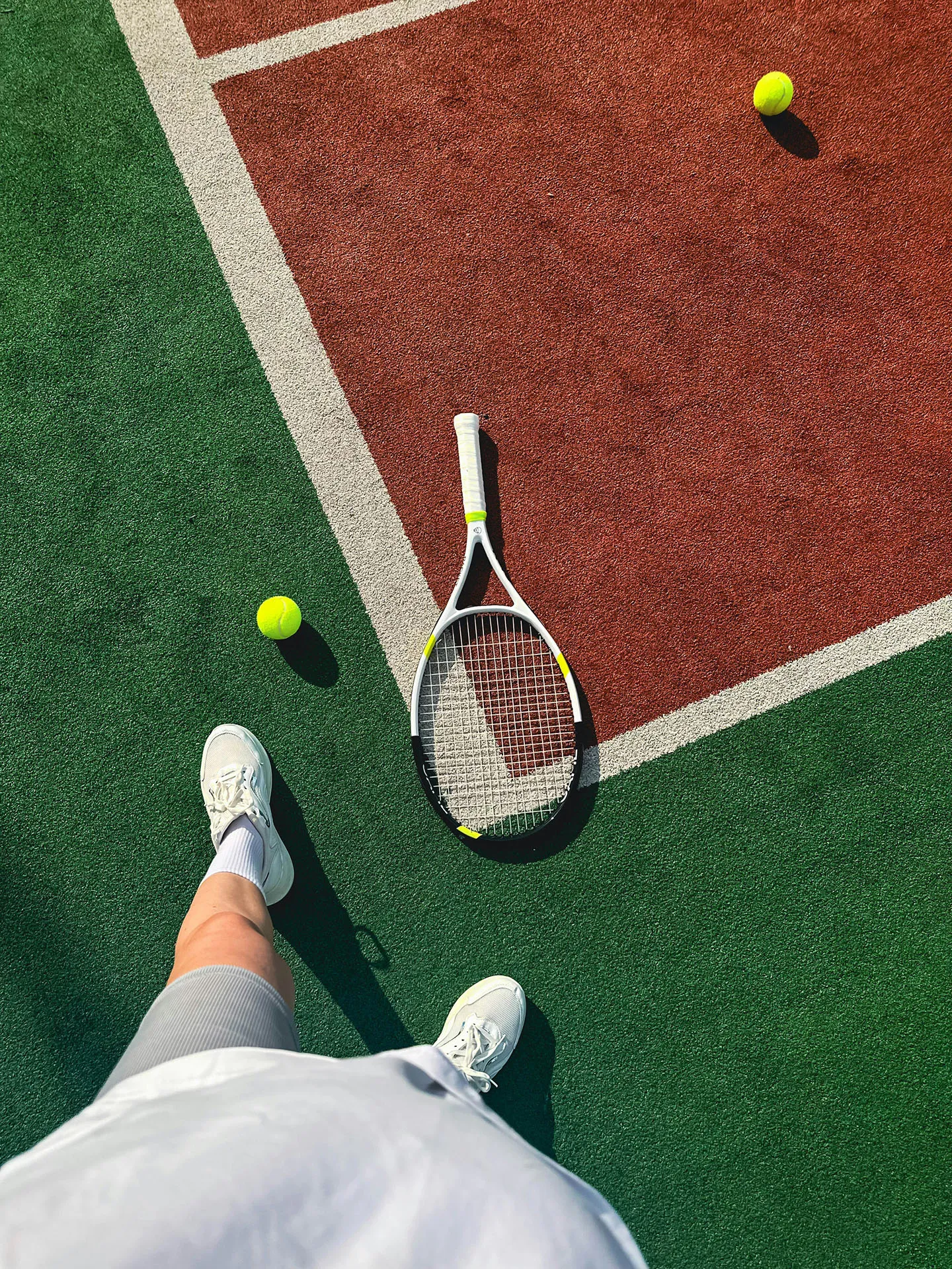 A person standing on a tennis court with a racket and two tennis balls on the ground.