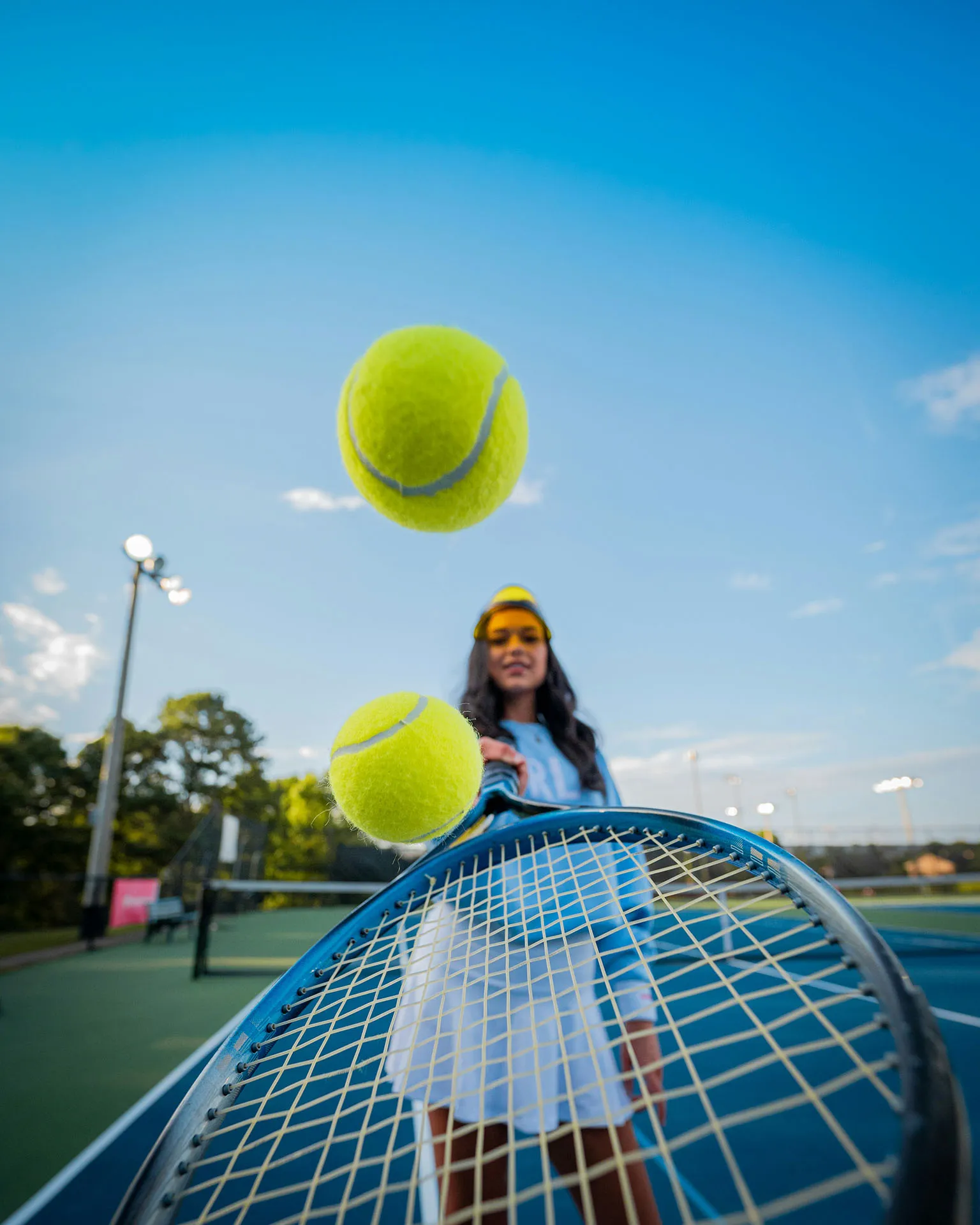 Woman holding tennis racket with two tennis balls in mid-air on a tennis court under a blue sky.