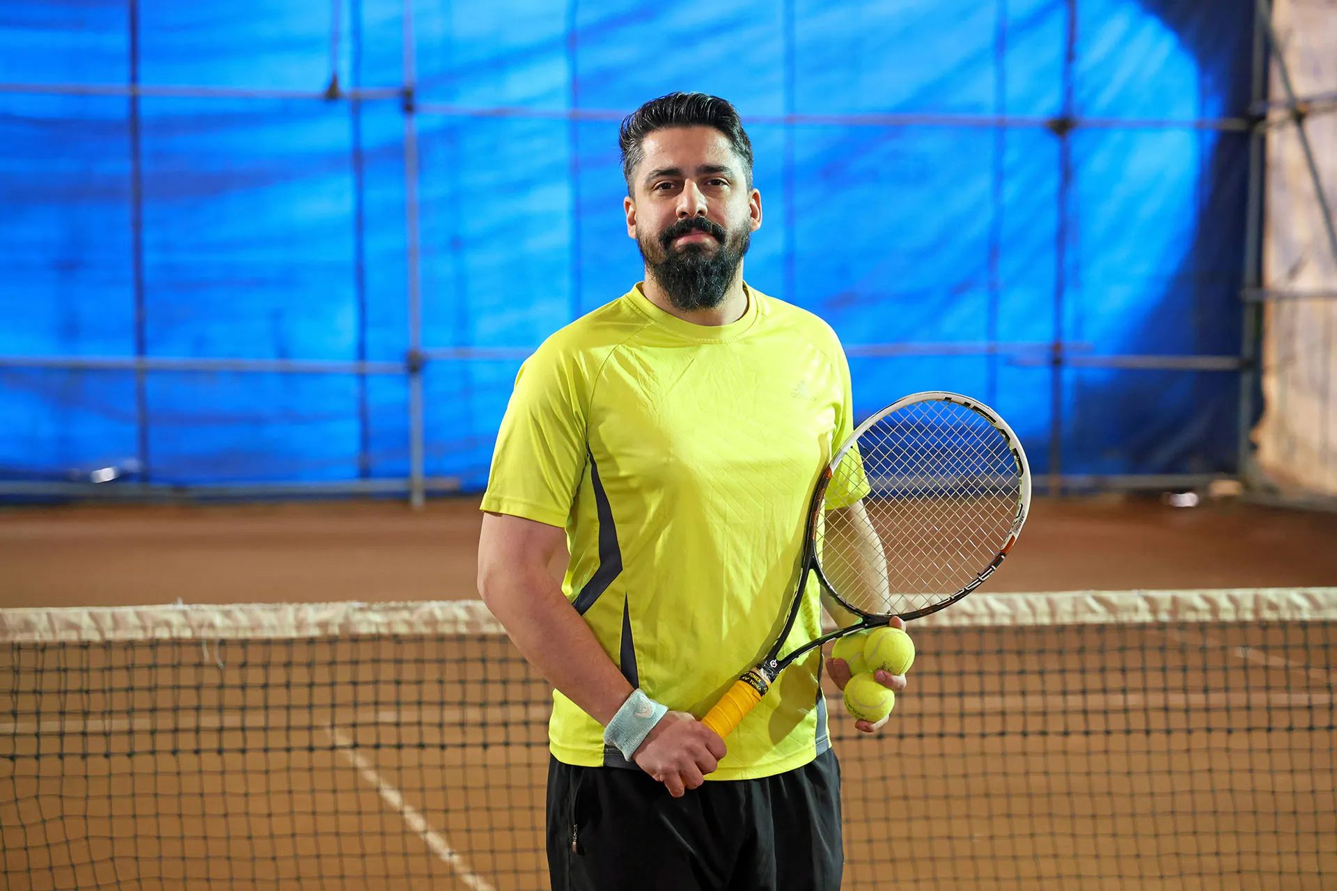 Man wearing a yellow sports shirt and black shorts holding a tennis racket and two tennis balls on an indoor tennis court.