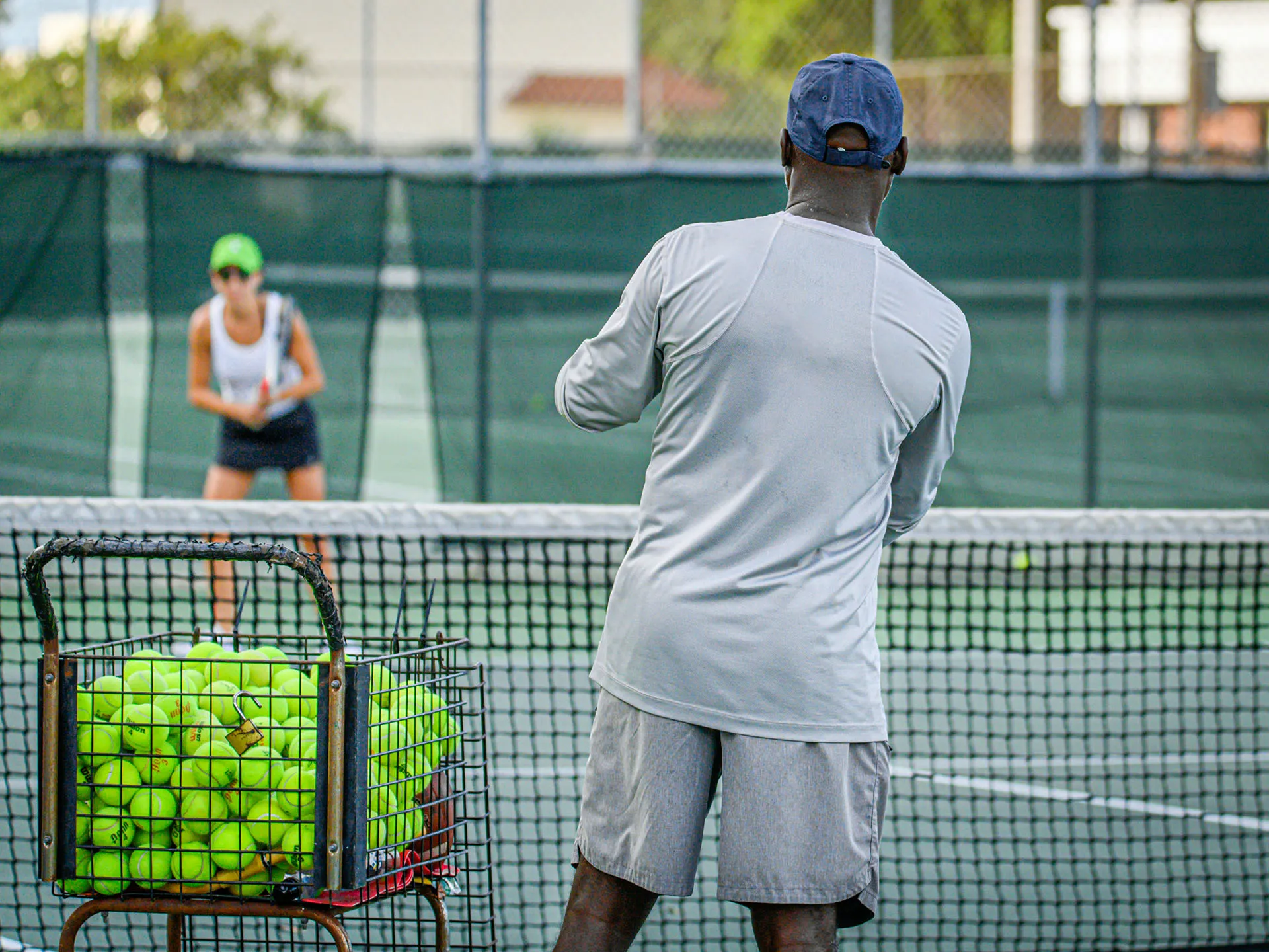 Tennis coach in gray shirt and cap hitting balls to a player wearing a green cap and white tank top on a tennis court with a basket of tennis balls.