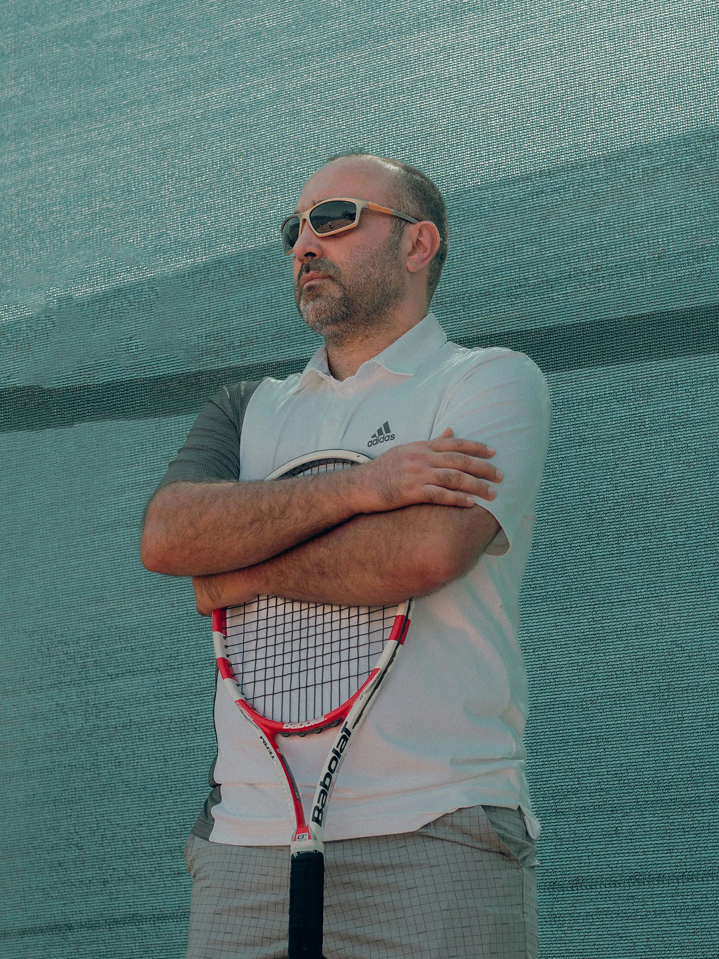 Man wearing sunglasses and a white Adidas shirt holding a Babolat tennis racket crossed over his chest, standing in front of a netted fence.