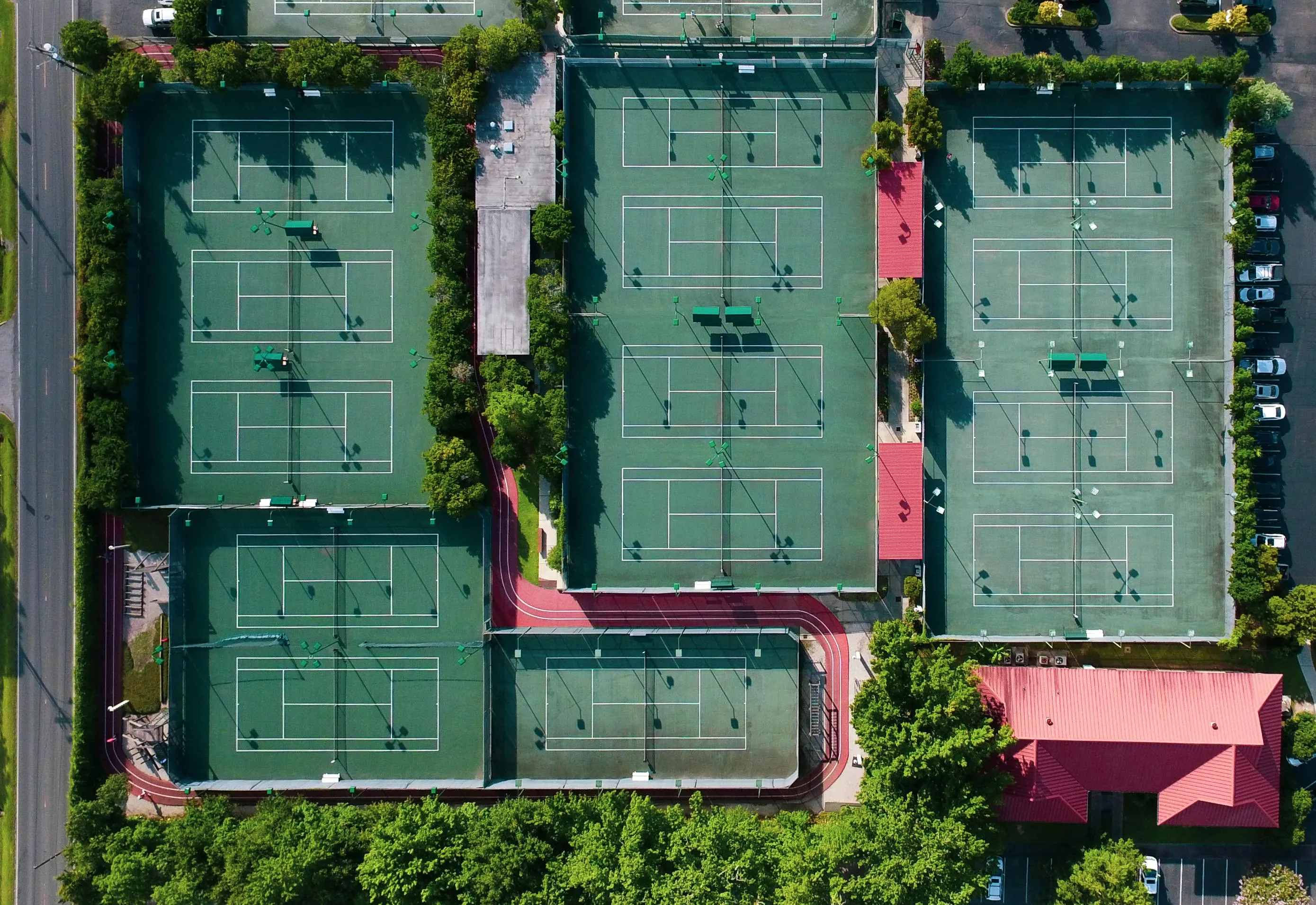 Aerial view of multiple green tennis courts with lighting and surrounding trees and buildings.