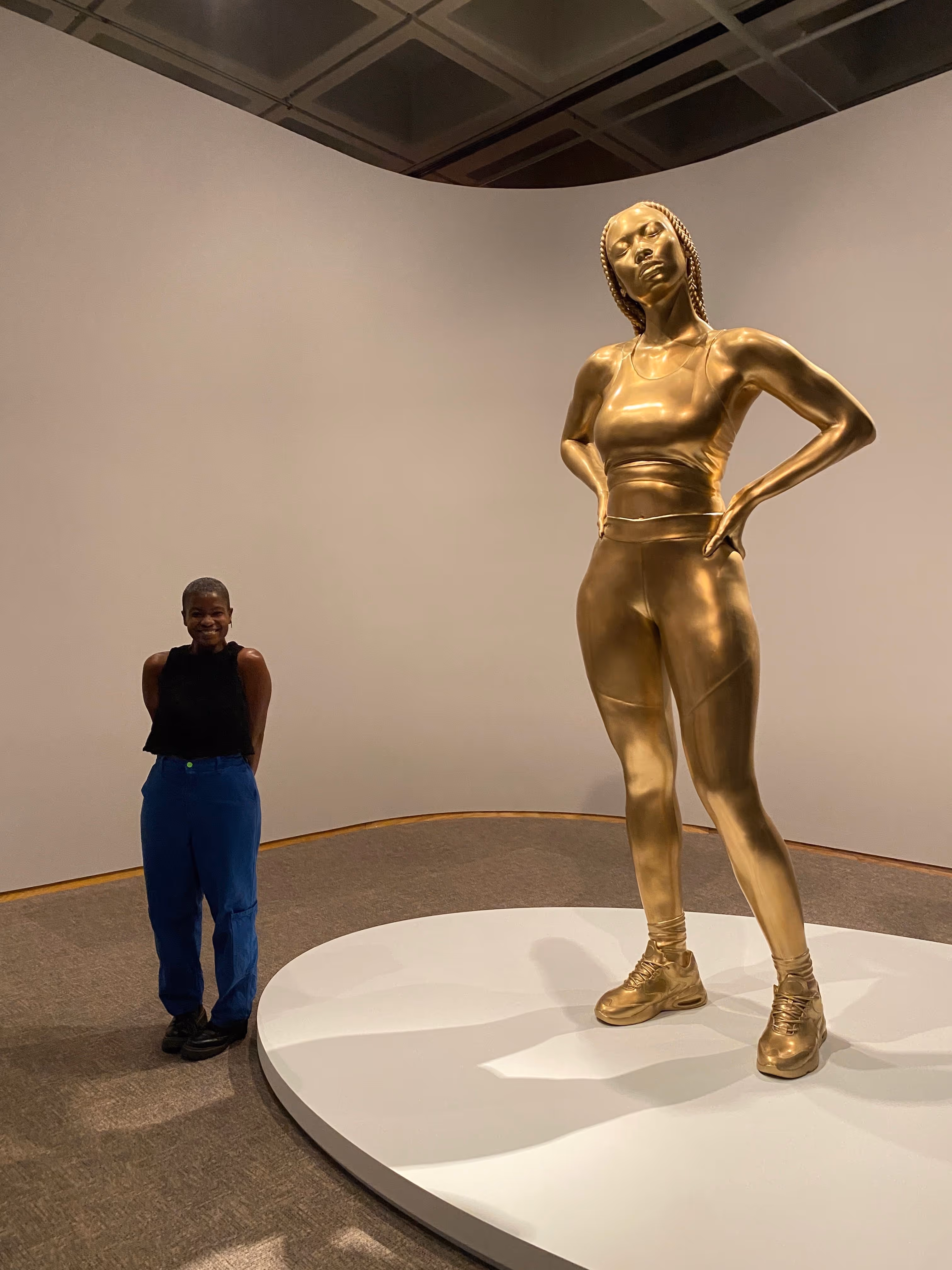 Niyia smiles while standing beside a towering gold sculpture of a woman in athletic wear—a sports bra, leggings, and sneakers—hands on hips, with long braids. The sculpture stands on a circular white platform in a gallery with white walls.