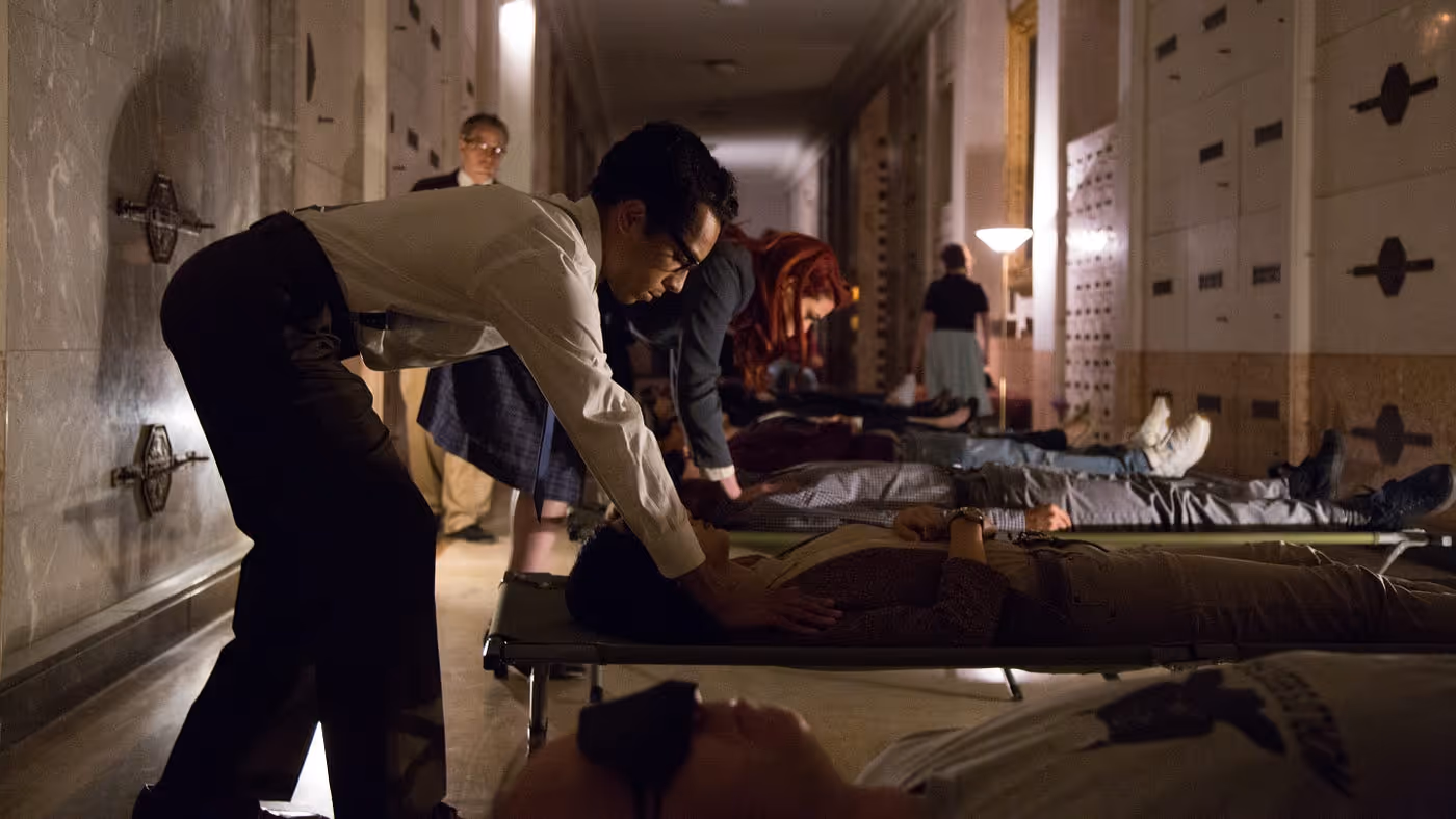 A dimly lit institutional hallway lined with safe deposit box walls. Multiple people lie on cots or the floor while standing figures attend to them. In the foreground, a man in a beige jacket leans over a prone figure on a cot. A floor lamp glows in the background, where additional figures move through the corridor.
