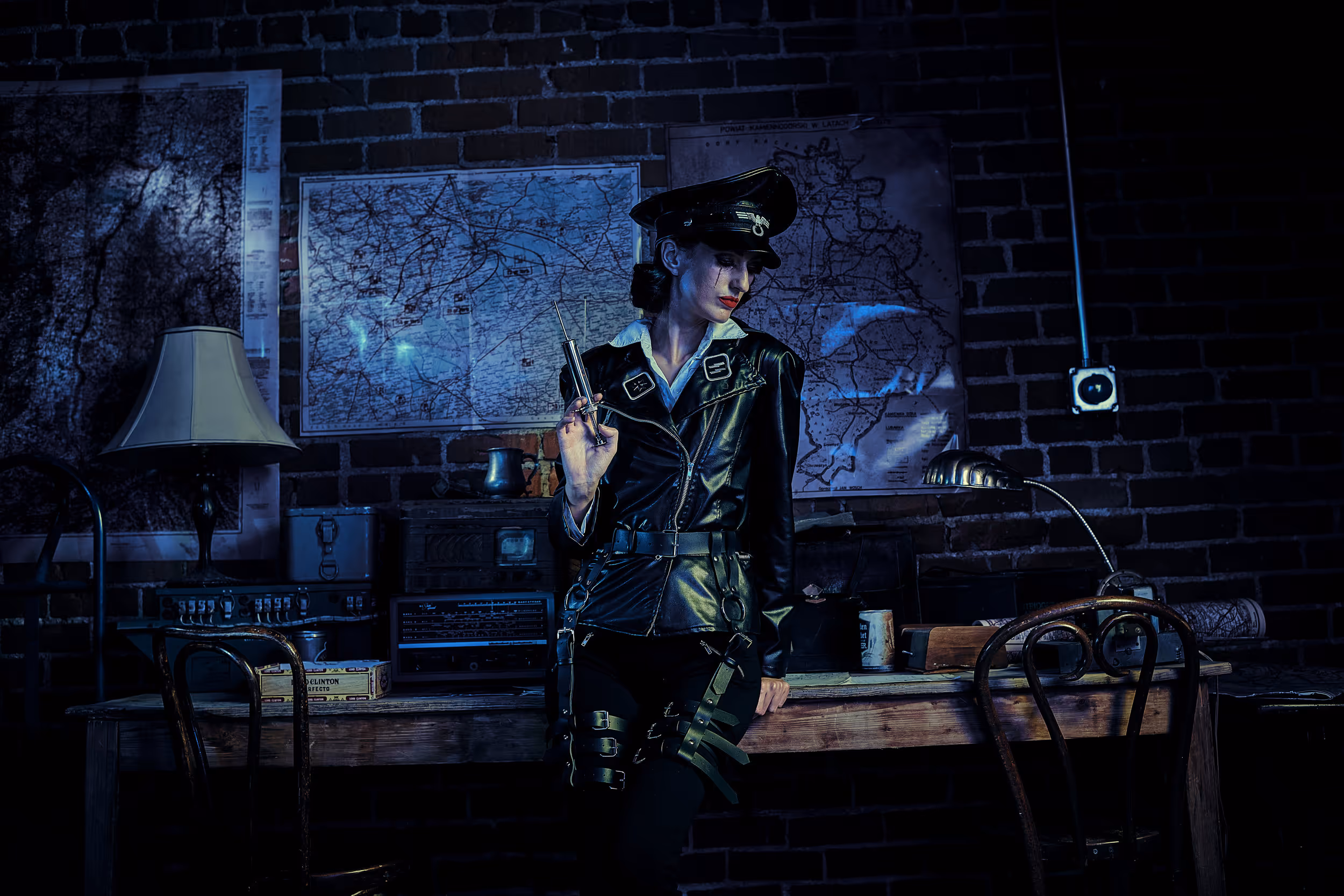 A woman in a black leather military-style uniform and peaked cap leans against a wooden desk in a darkly lit brick room, holding a syringe. Behind her, vintage maps are pinned to the wall alongside old radio equipment, a table lamp, and a cigar box. The scene is lit in deep blue tones.