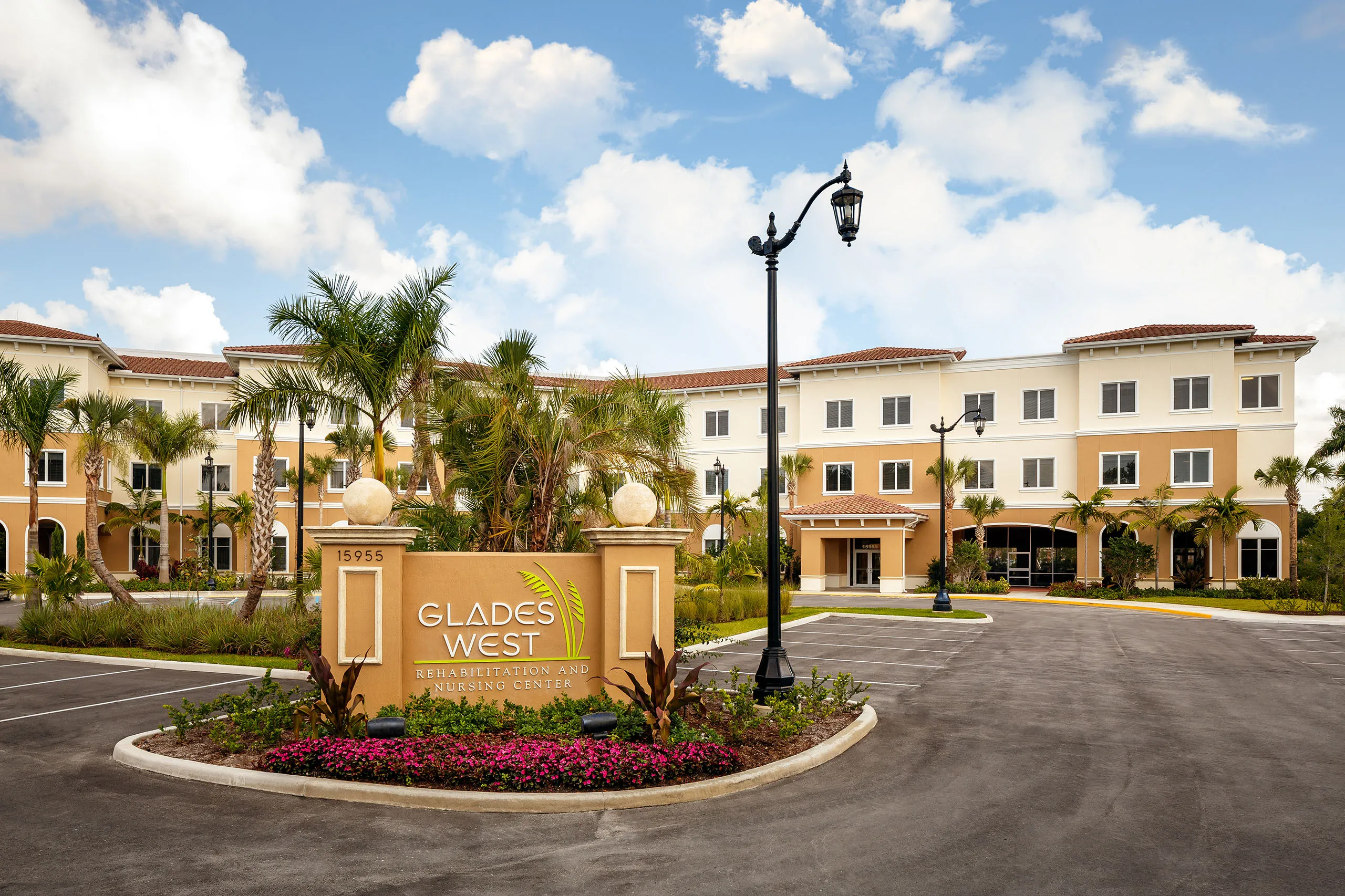 Front view of Glades West Rehabilitation and Nursing Center with palm trees, flower beds, and parking area under a partly cloudy sky.