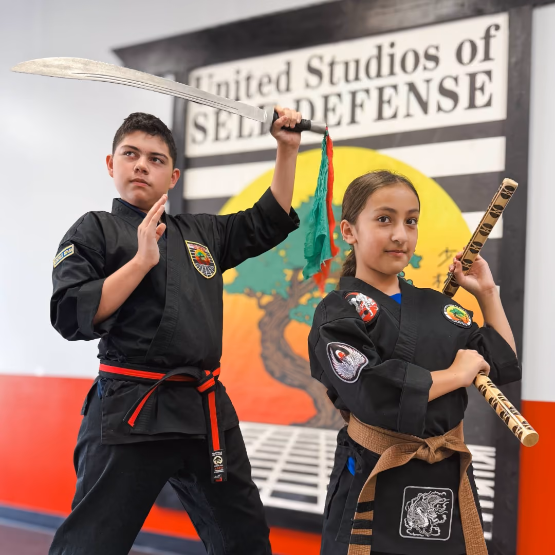 Two young martial artists in black uniforms posing with a sword and wooden sticks inside a dojo.