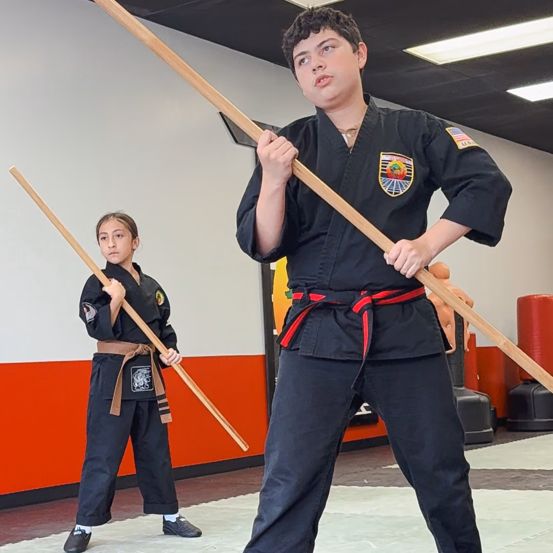 Two martial arts students in black uniforms holding wooden bo staffs in a training room.