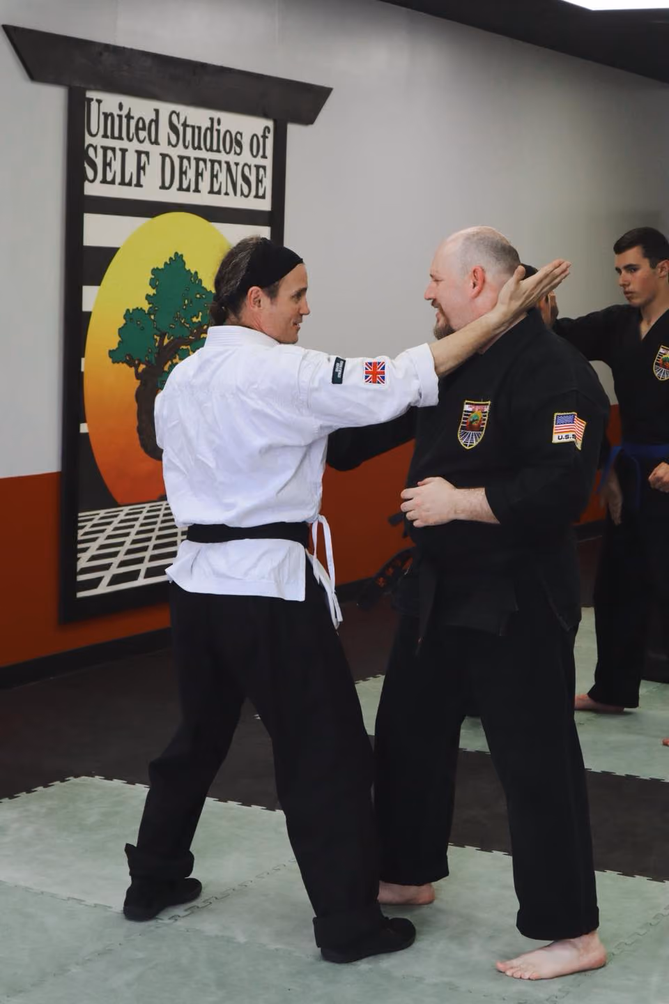Two martial artists sparring in black and white uniforms in a self-defense studio with a tree logo.