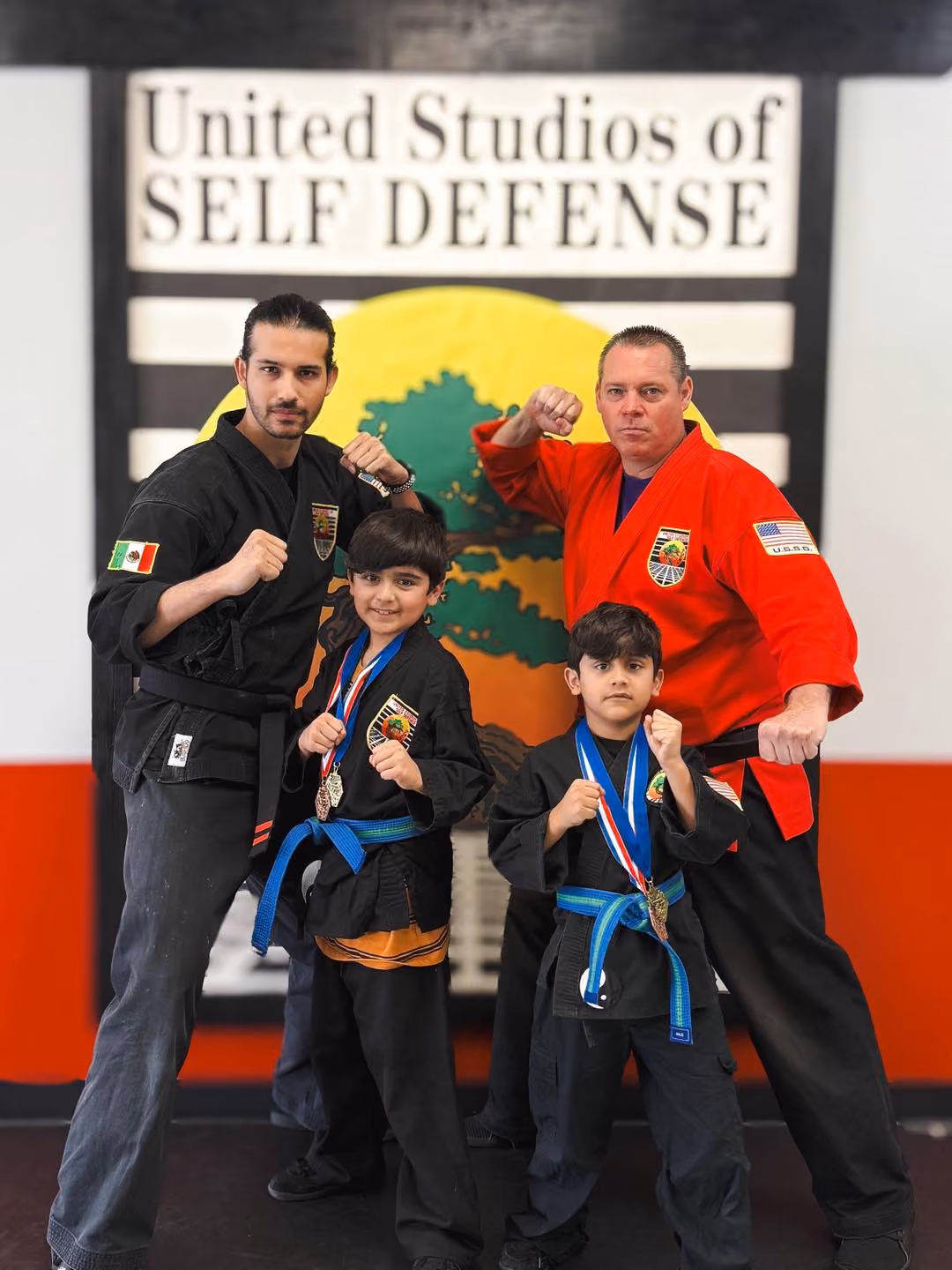 Two men and two boys in martial arts uniforms posing with fists raised in front of USSD studio sign.
