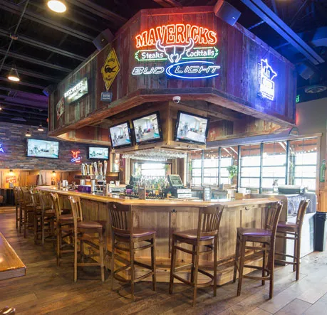 Wooden bar with stools arranged around it, neon signs reading Mavericks Steaks Cocktails, and multiple TV screens hanging above.