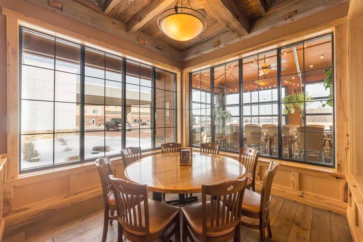 Cozy dining area with a round wooden table surrounded by eight chairs, large windows with black grid frames, and warm wooden walls and ceiling beams.