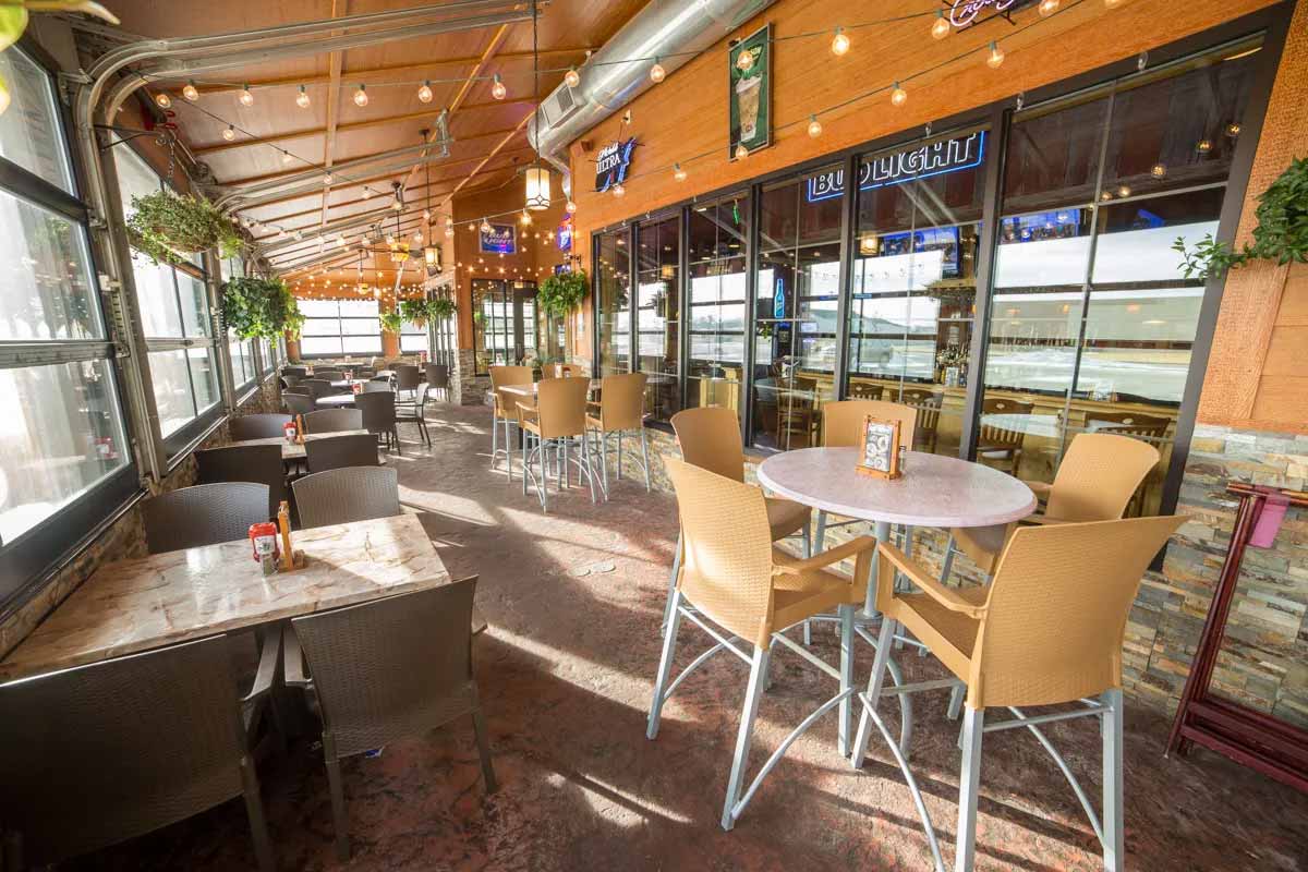 Sunlit outdoor restaurant patio with tables and chairs, hanging plants, and string lights along a wooden ceiling.