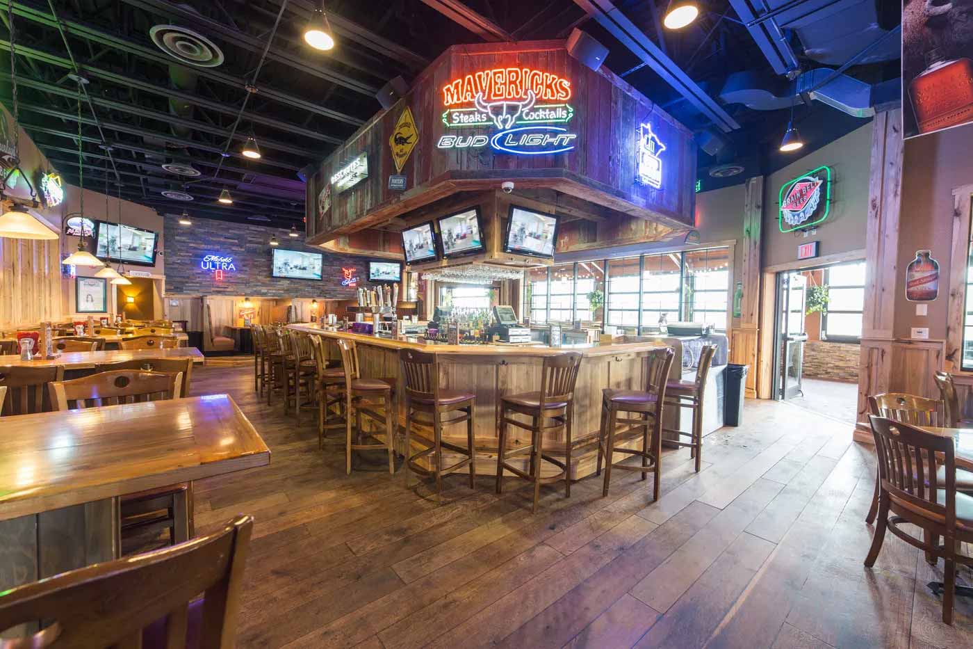 Rustic bar interior with wooden floors and chairs, central hexagonal bar counter with multiple stools, neon signs, and several TV screens.