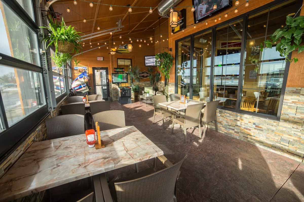 Bright indoor patio area with marble-top tables, wicker chairs, hanging plants, string lights, and TVs mounted on walls.