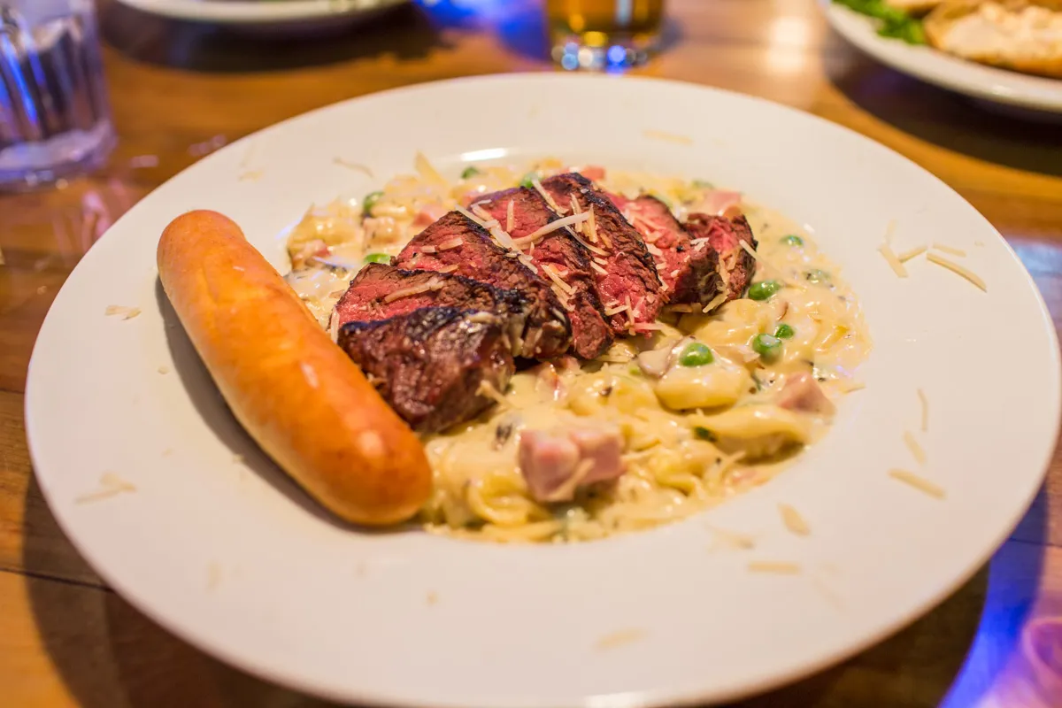 Plate with creamy pasta containing peas and ham, topped with sliced grilled steak and a breadstick on the side.