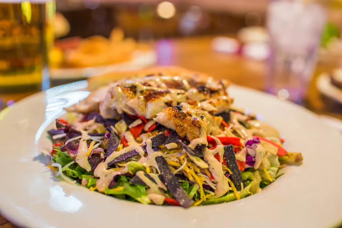 Grilled chicken breast atop mixed greens salad with shredded cheese, purple and red tortilla strips, drizzled with creamy dressing on a white plate.