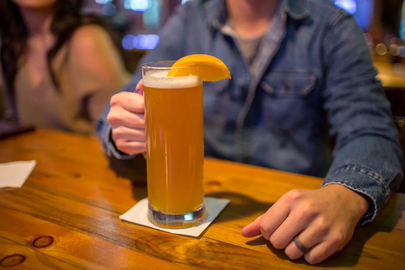 Person in denim jacket holding a tall glass of beer garnished with an orange slice on a wooden table.
