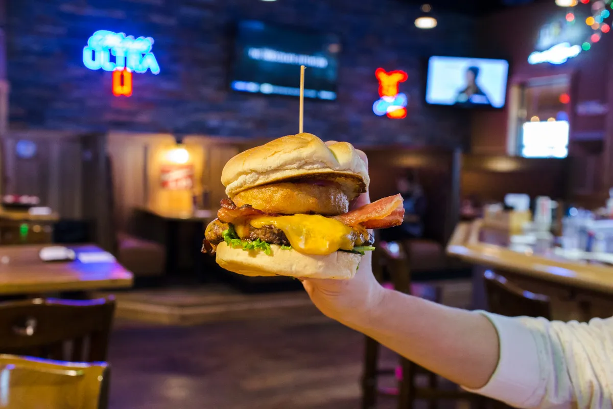 Hand holding a cheeseburger with bacon, cheddar cheese, lettuce, and an onion ring inside a restaurant setting.