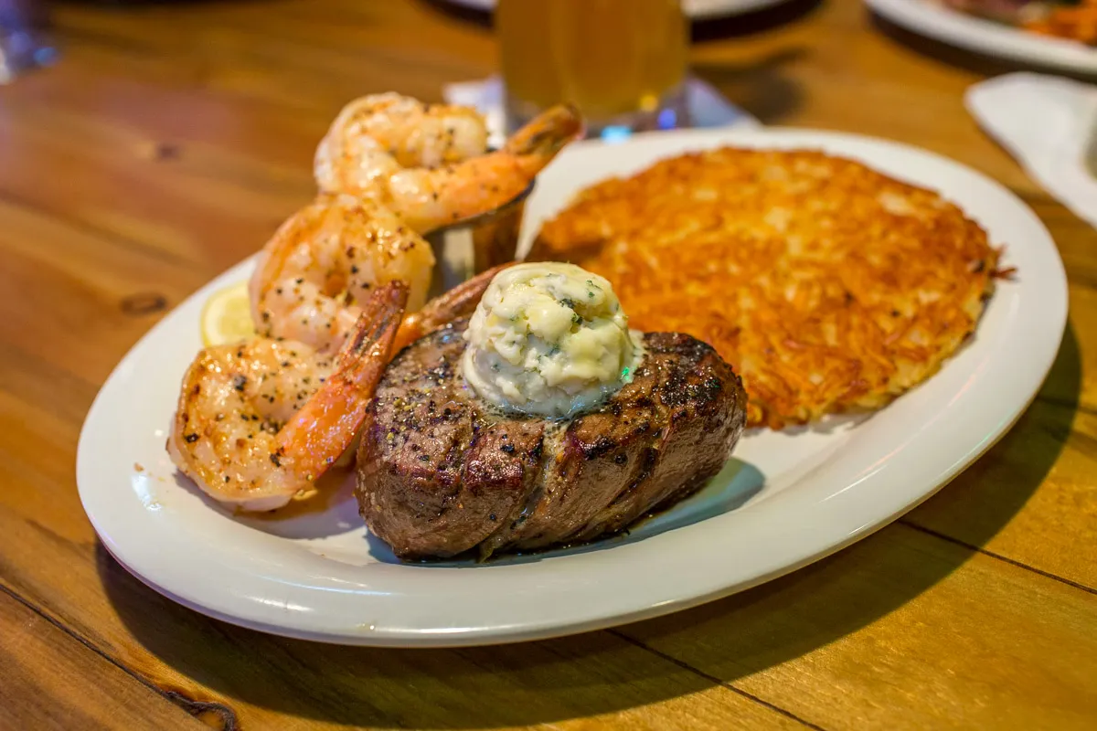 Plate with grilled shrimp, a filet mignon topped with herb butter, and a large crispy hash brown cake on a wooden table.