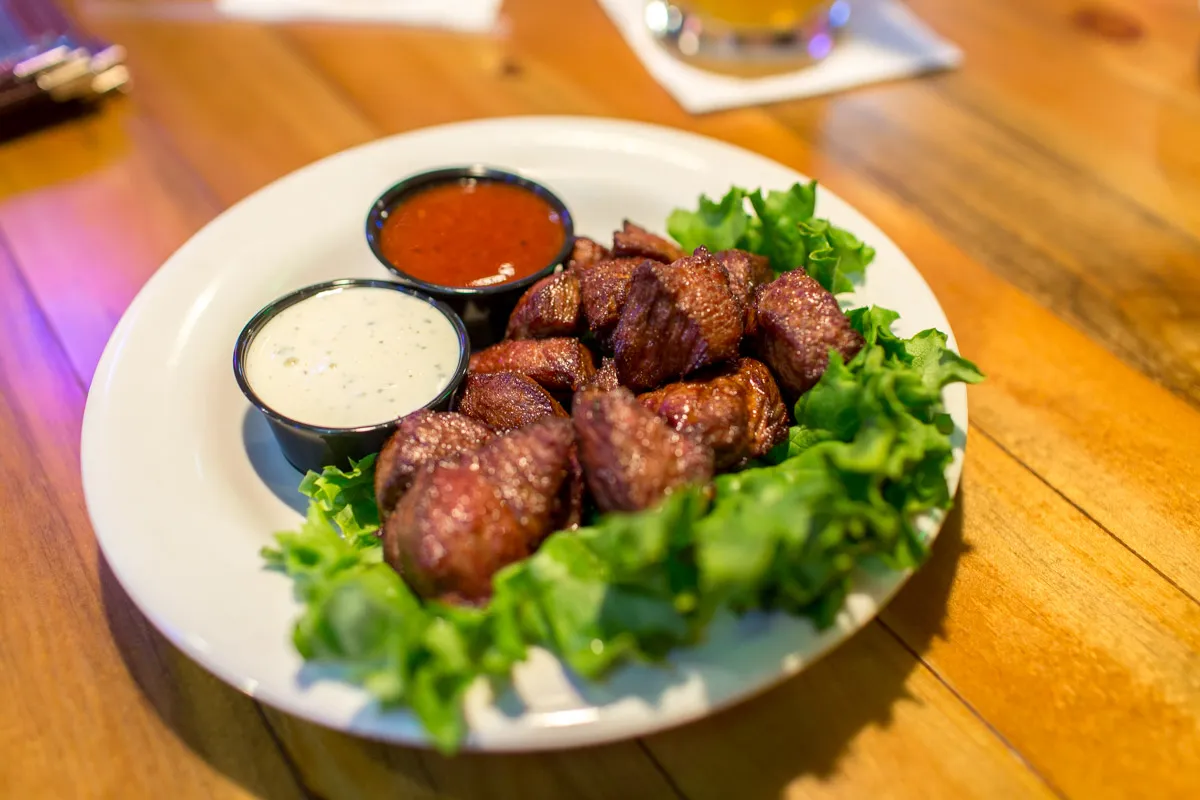 Plate of grilled steak bites served on lettuce with two dipping sauces, one creamy white and one red.