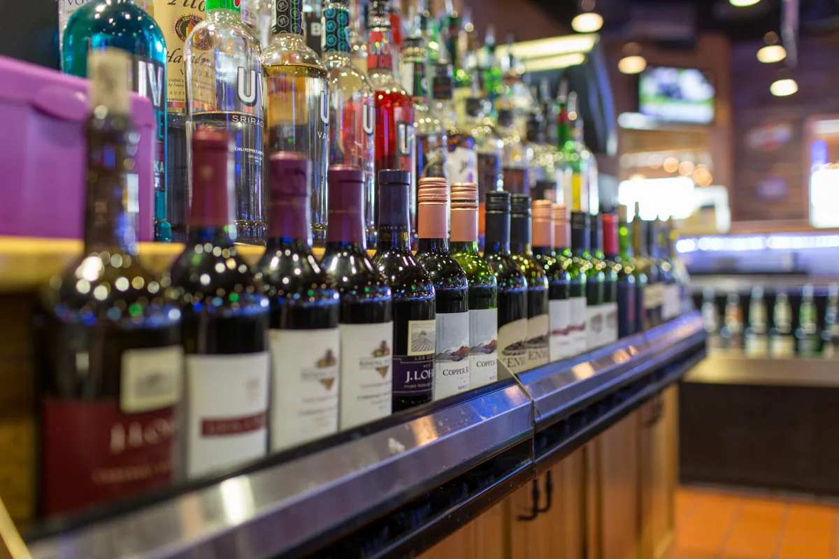 Rows of wine and liquor bottles lined up on a bar counter with a blurred restaurant interior in the background.